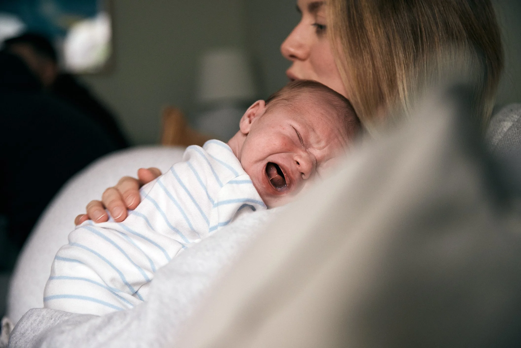 A woman holding a crying newborn baby close to her chest, with the baby's face pressed against her face, in an indoor setting.