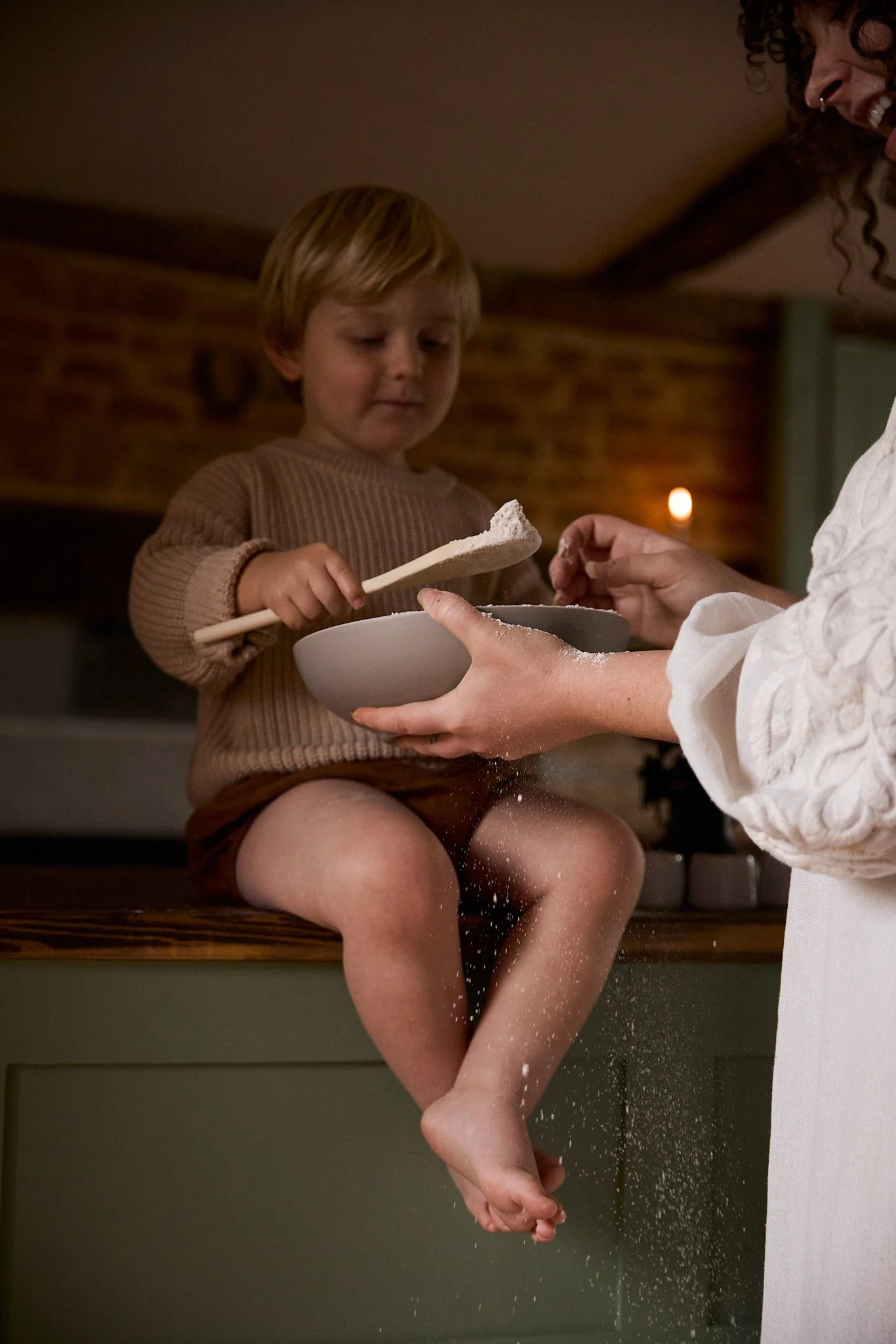 A young boy sitting on a kitchen counter with powder on his legs, being fed flour by a woman, possibly his mother, in a cozy kitchen setting.