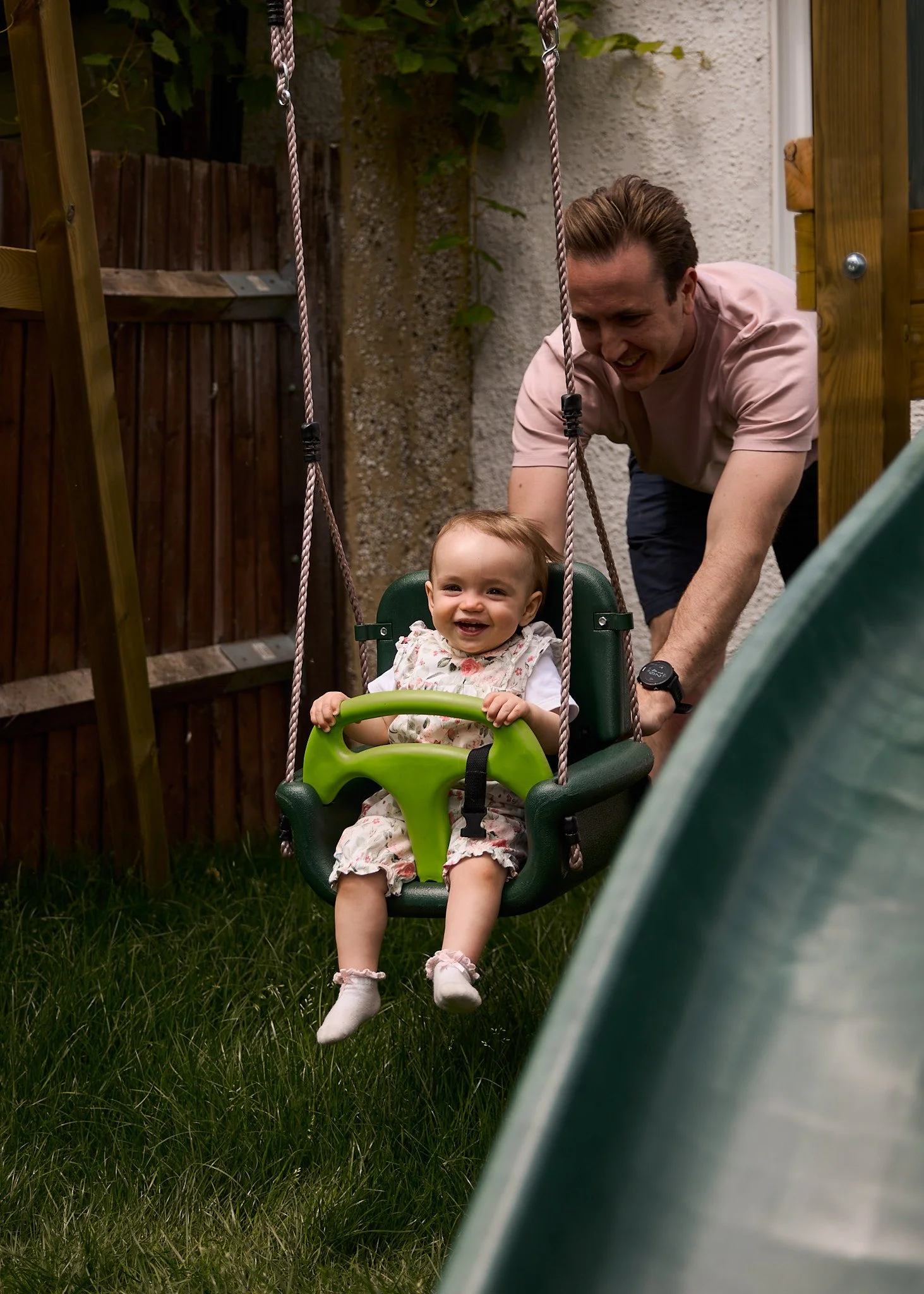 A young girl happily swinging on a backyard swing with a man pushing her, next to a slide, in a grassy yard enclosed by a wooden fence.