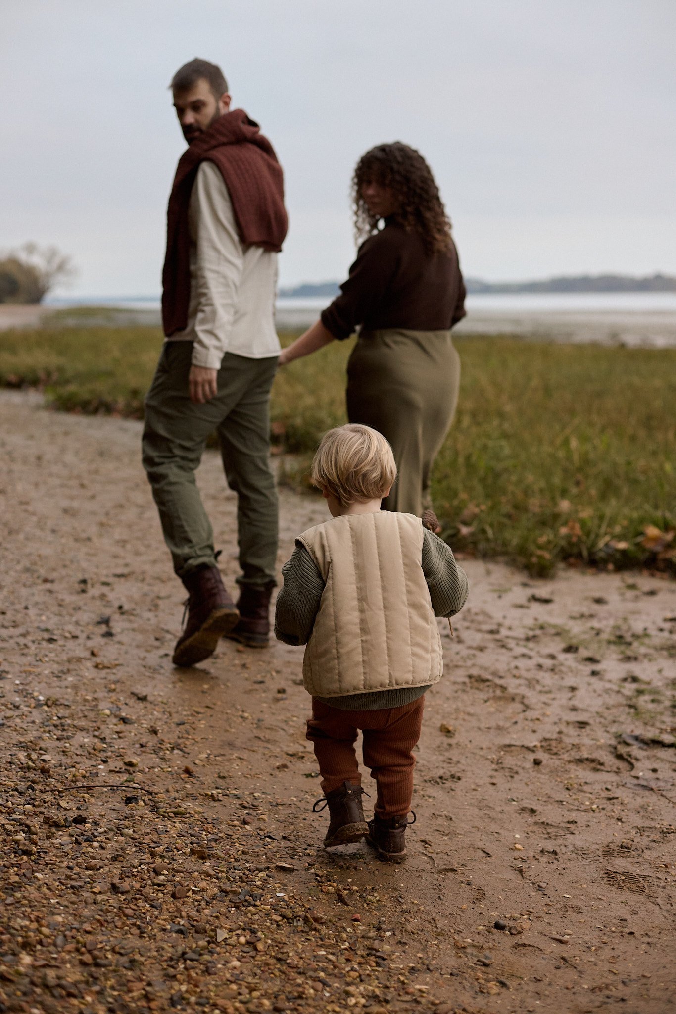 A family walking along a sandy, rocky trail near a grassy area by water, with a man, woman, and young child seen from behind.