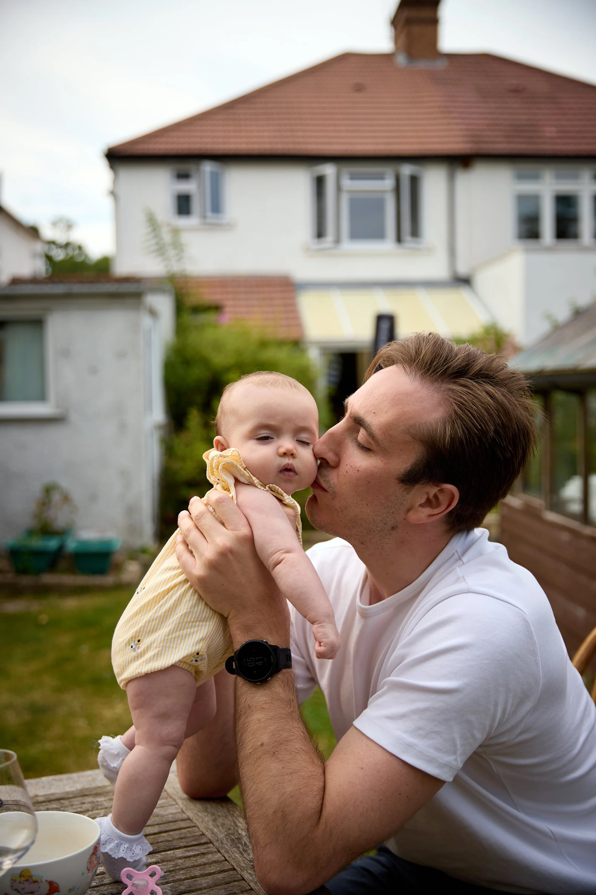 A man holding a young baby girl outdoors in a backyard, about to kiss her cheek, with houses and trees in the background.