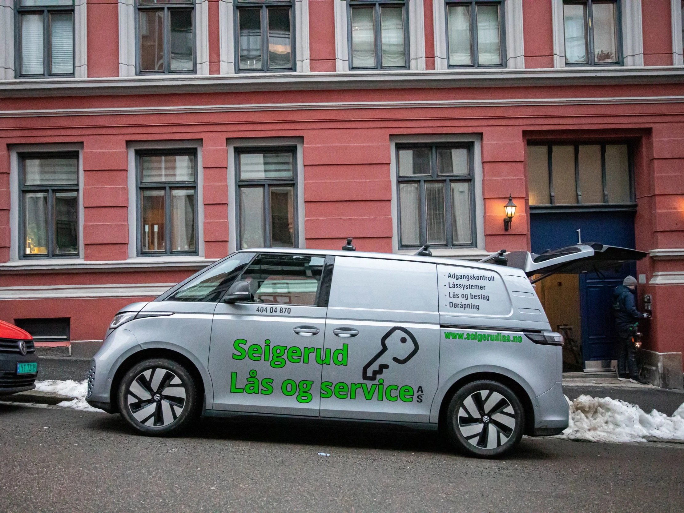 Fotografi av en bygate med biler parkert langs gaten foran en rød bygning med flere vinduer. På en av bilene er det en reklame med grønne bokstaver som sier "Seigerud lås & service", og tekst på siden av bilen annonserer lås- og servicesystemer. Det er snø på bakken.
