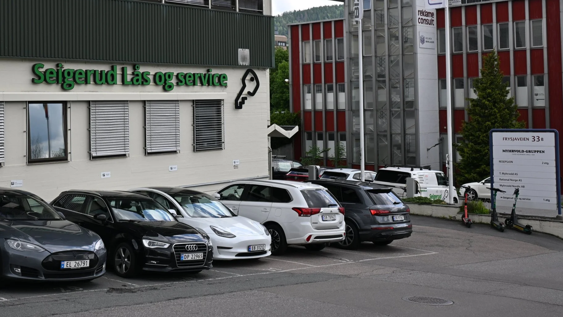 Parkering foran en bygning med skiltet 'Seigerud Lås og service', en annen bygning i bakgrunnen har skiltet 'reklaаме consult', flere biler parkert på parkeringsplassen, scooter ved siden av en informasjonstavle.