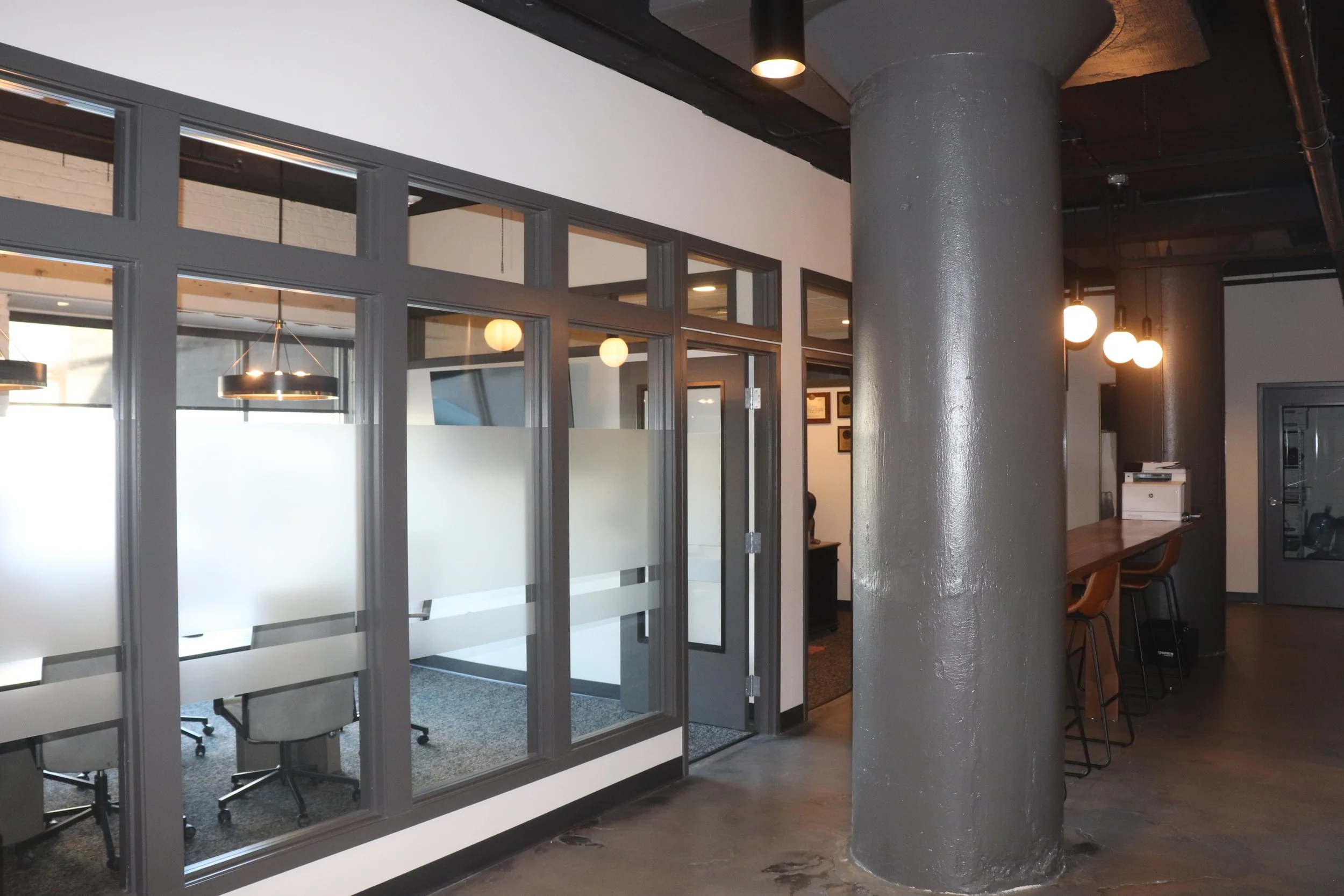 Modern office interior with glass-walled conference room, large gray support pillar, and a wooden bar-height counter with chairs
