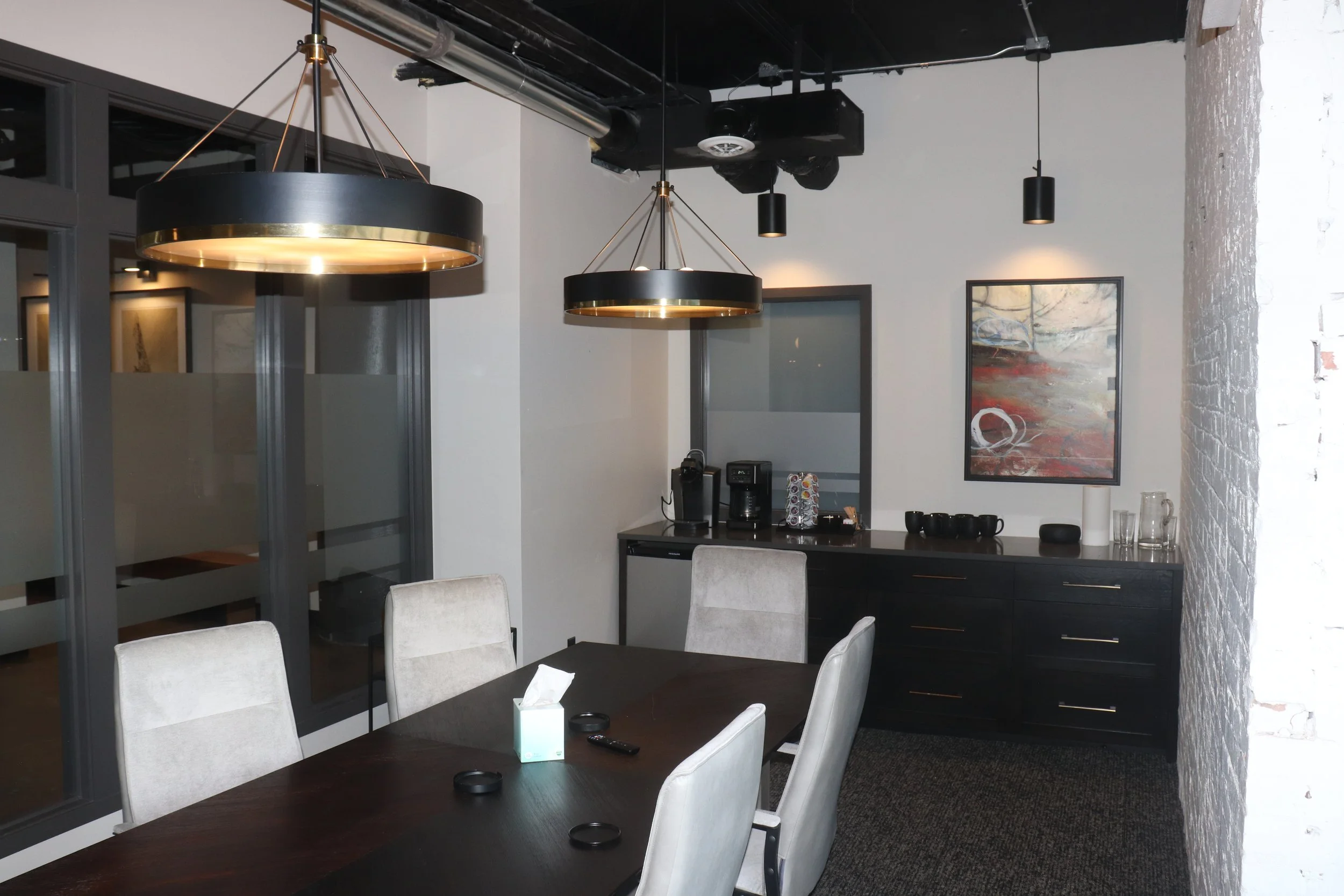 A modern dining room with a dark wood table and six beige upholstered chairs. Two large black and gold pendant lights hang over the table. There's a black sideboard with cups, a faucet, and decorative items, and a coffee station with a Keurig and a K