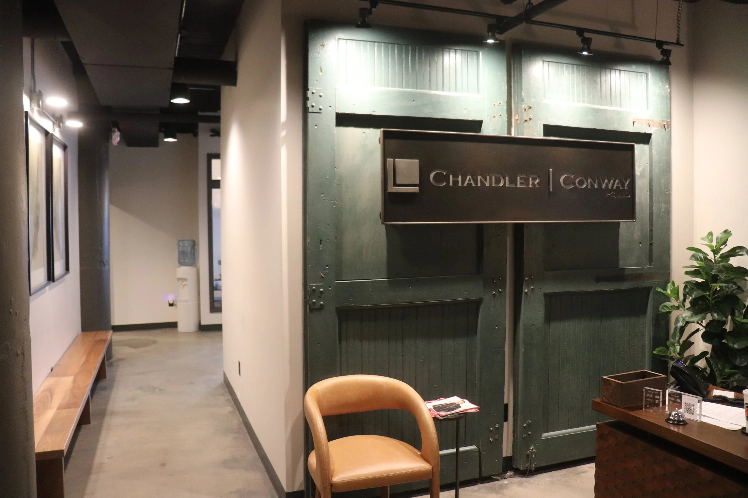 Interior of an office or lobby area with a green door, a leather chair, a small table with magazines, and a reception desk with a plant and office items. The door has a sign that says 'Chandler Conway'.
