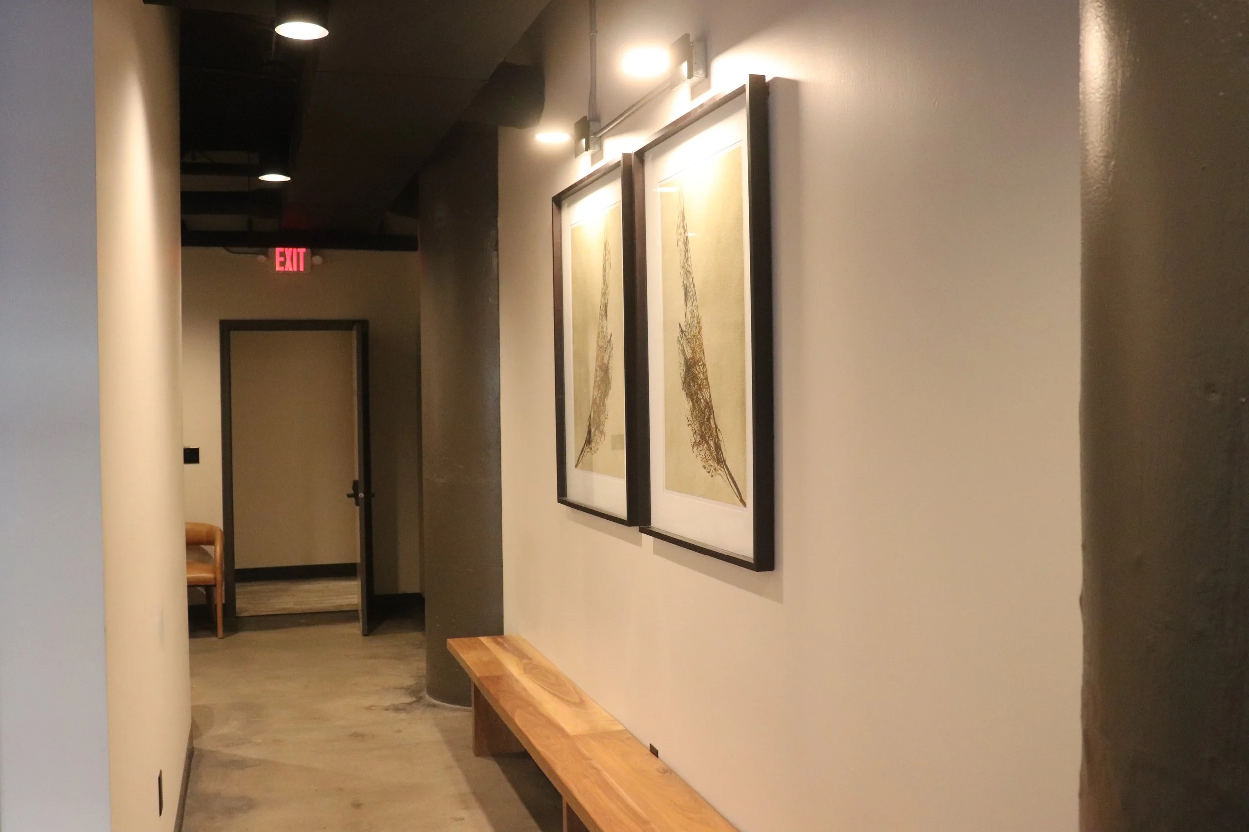 A hallway with two framed abstract art pieces on the right wall. There's a wooden bench below the artwork. In the background, an open door, an illuminated exit sign, and a corner with another chair are visible. The floor is concrete, and the walls ar