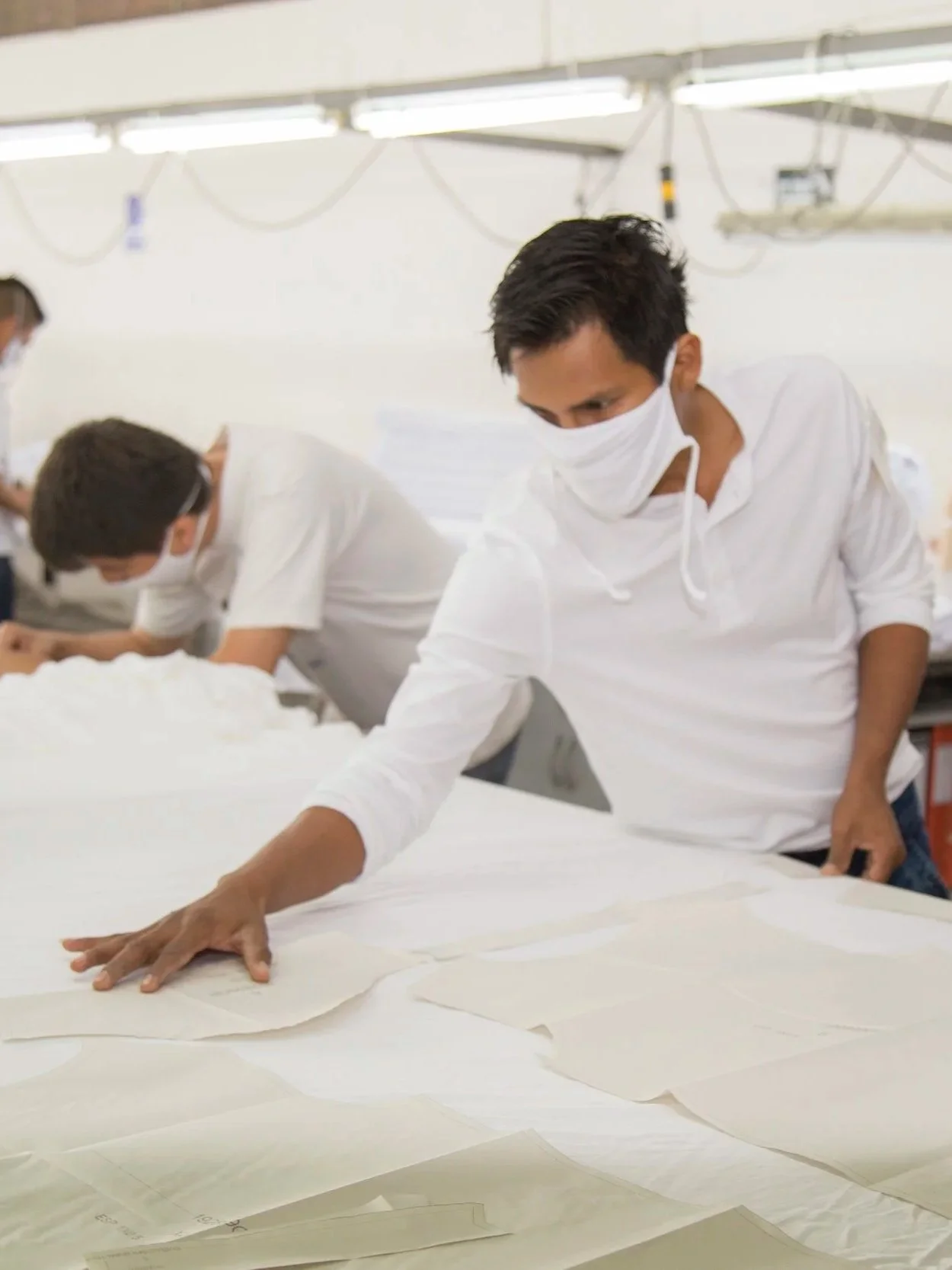People working in a textile factory, wearing masks, arranging fabric patterns on a large table under bright lights.