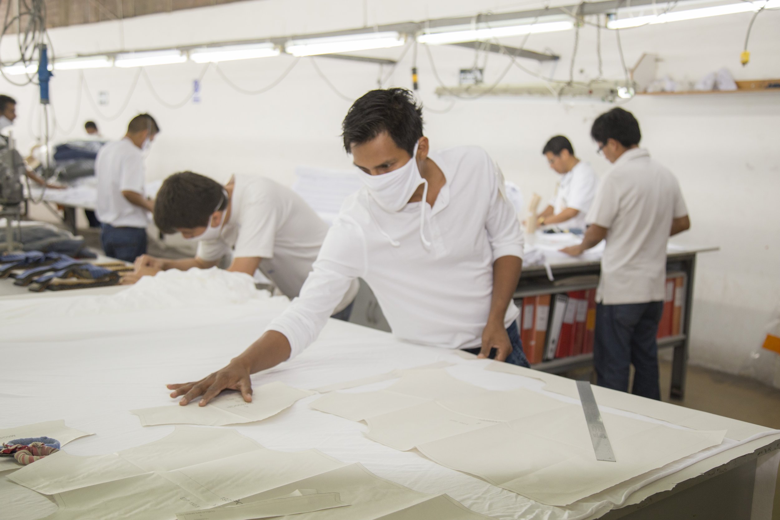 People working in a textile factory, wearing masks, arranging fabric patterns on a large table under bright lights.