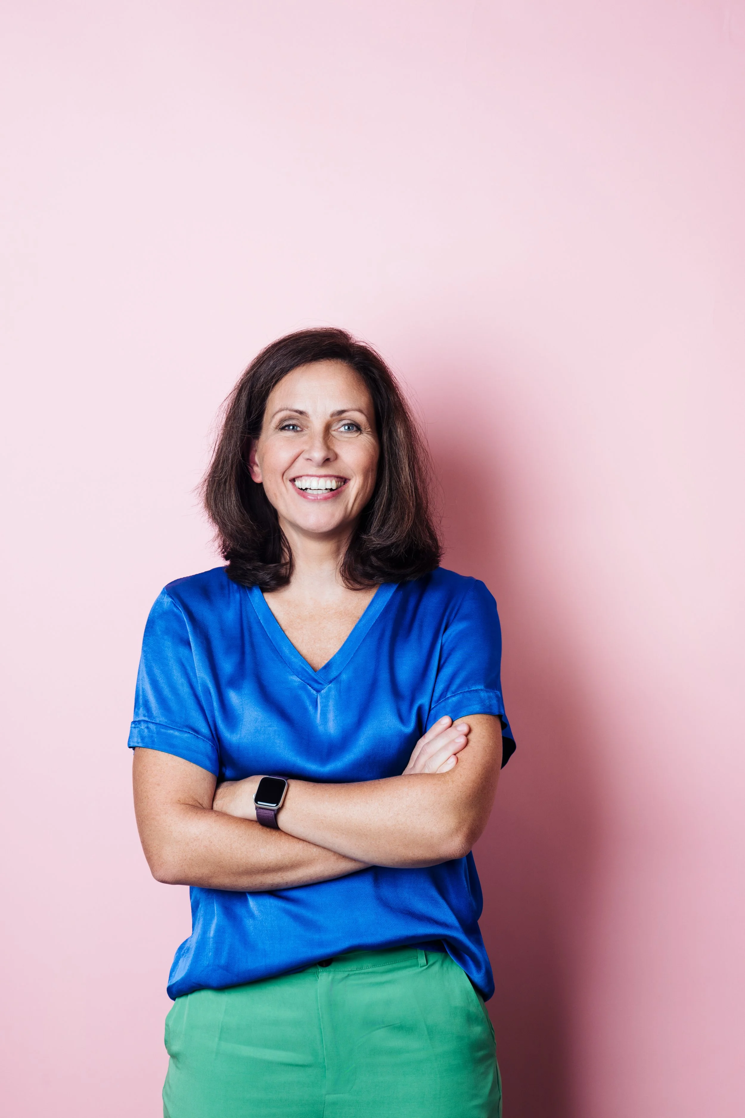 Eine smiling Frau mit braunen, schulterlangen Haaren, trägt eine blaue Bluse, eine grüne Hose und eine schwarze Smartwatch am Handgelenk, steht vor einem rosa Hintergrund.