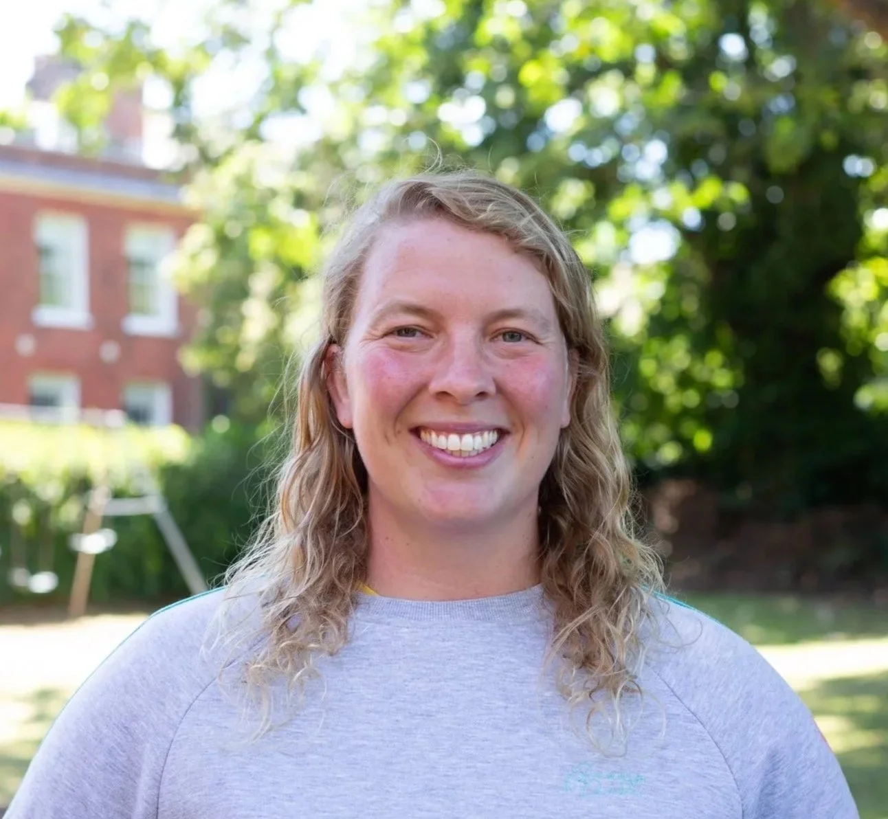 A smiling woman with curly, shoulder-length hair standing outdoors in front of a brick wall.