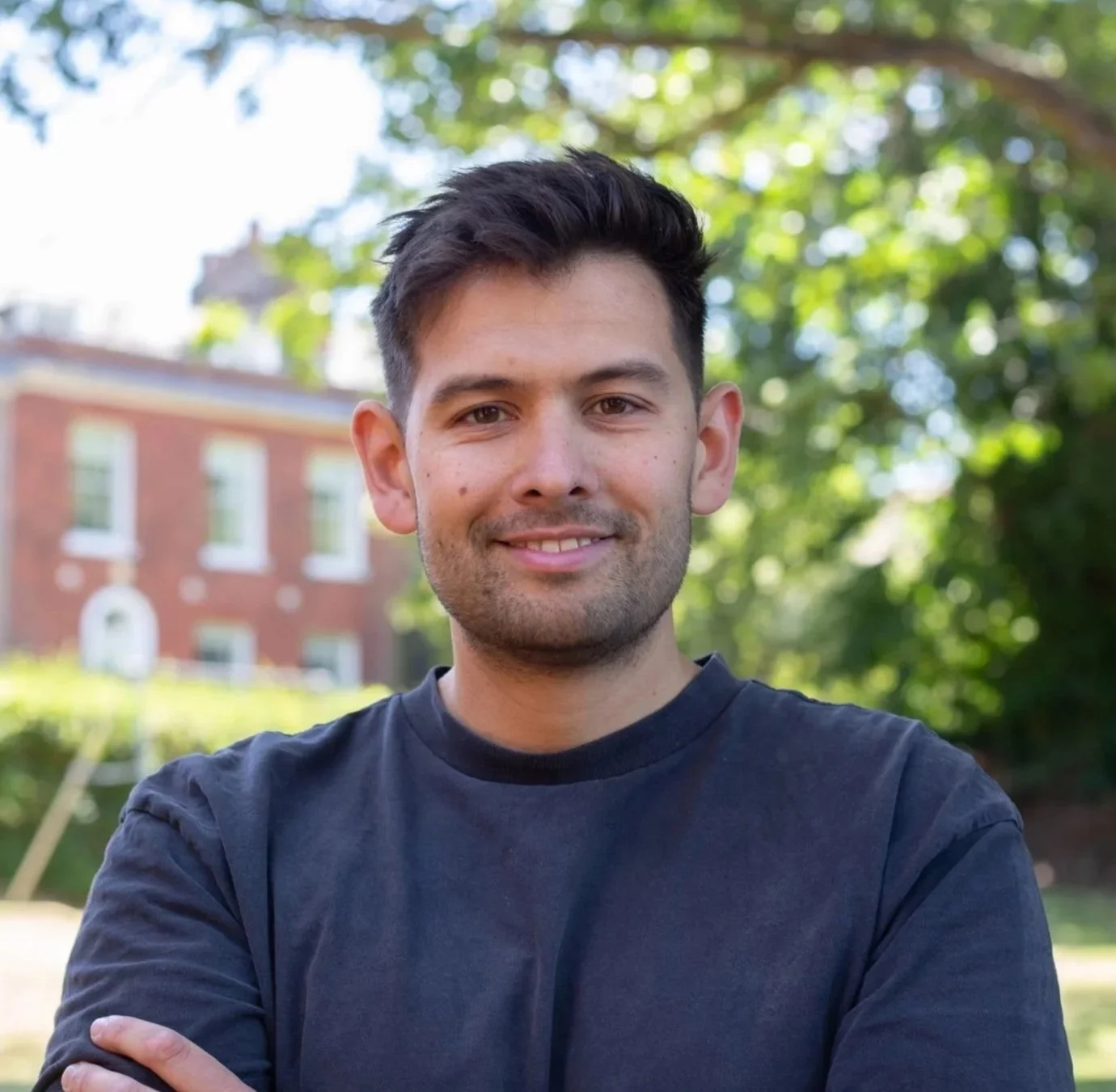 A smiling young man with short dark hair, wearing a black button-up shirt over a white T-shirt, standing outdoors in front of a brick wall and greenery.