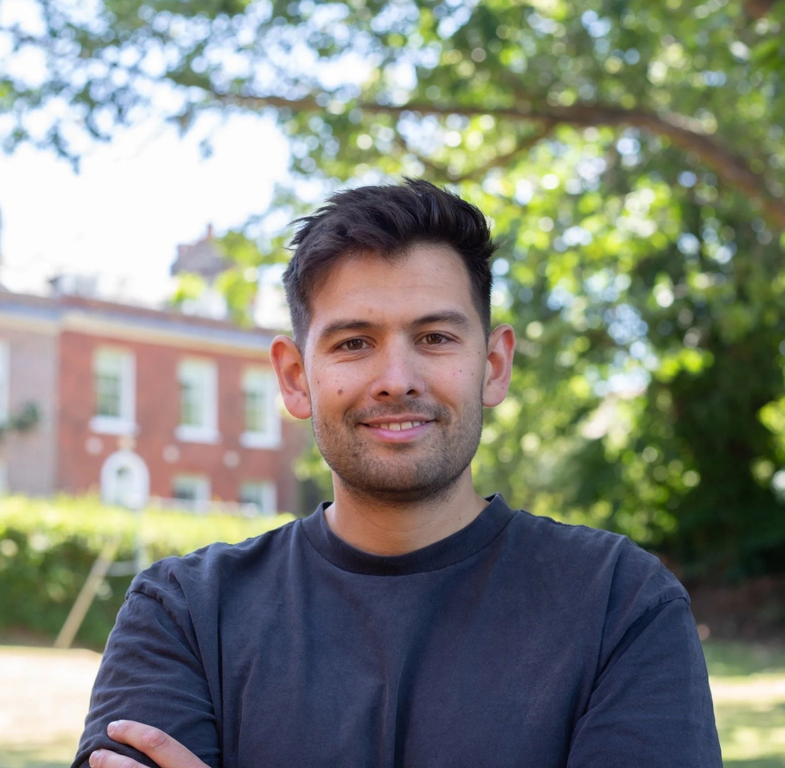 A smiling young man with short dark hair, wearing a black button-up shirt over a white T-shirt, standing outdoors in front of a brick wall and greenery.
