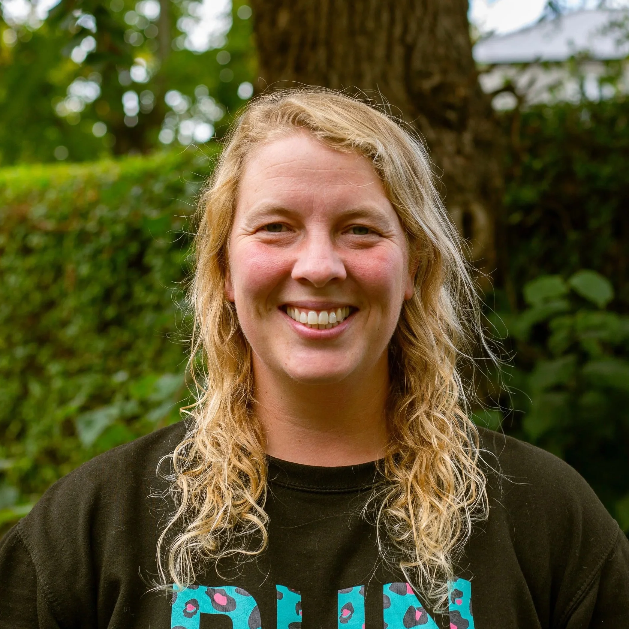 A smiling woman with curly, shoulder-length hair standing outdoors in front of a brick wall.