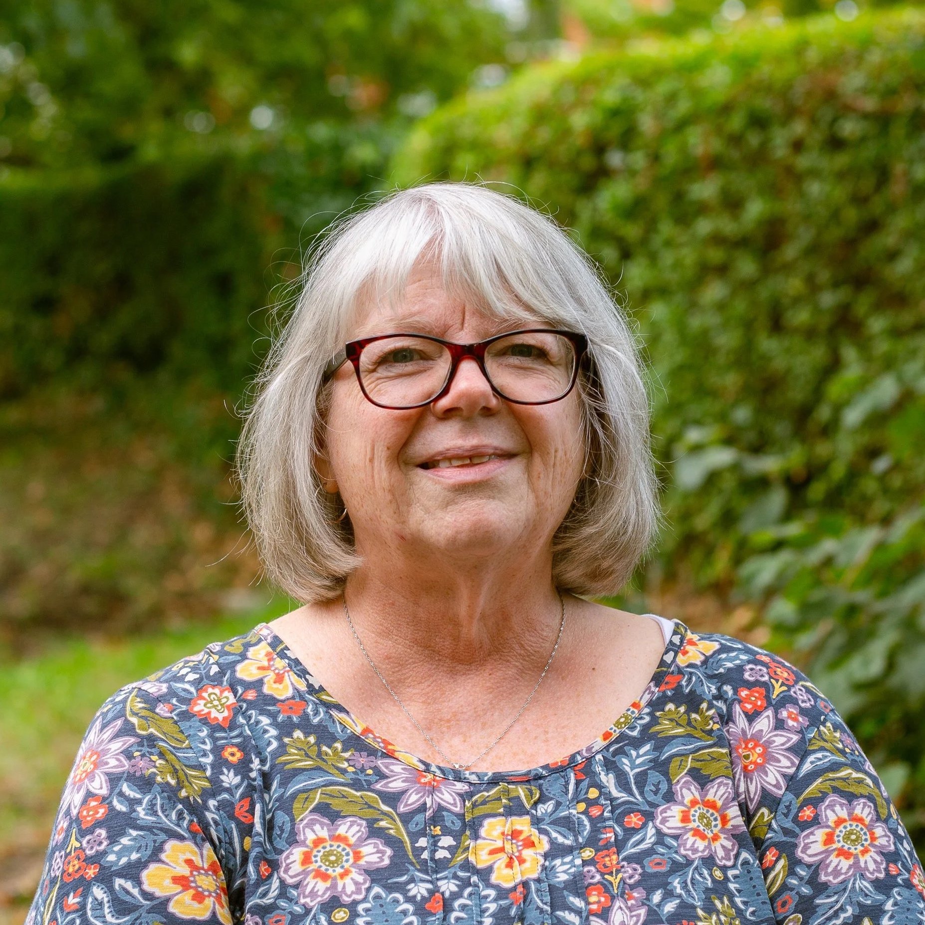 Smiling elderly woman with glasses and long gray hair outdoors, wearing a pink cardigan over a black top and a cross necklace, standing in front of green foliage.