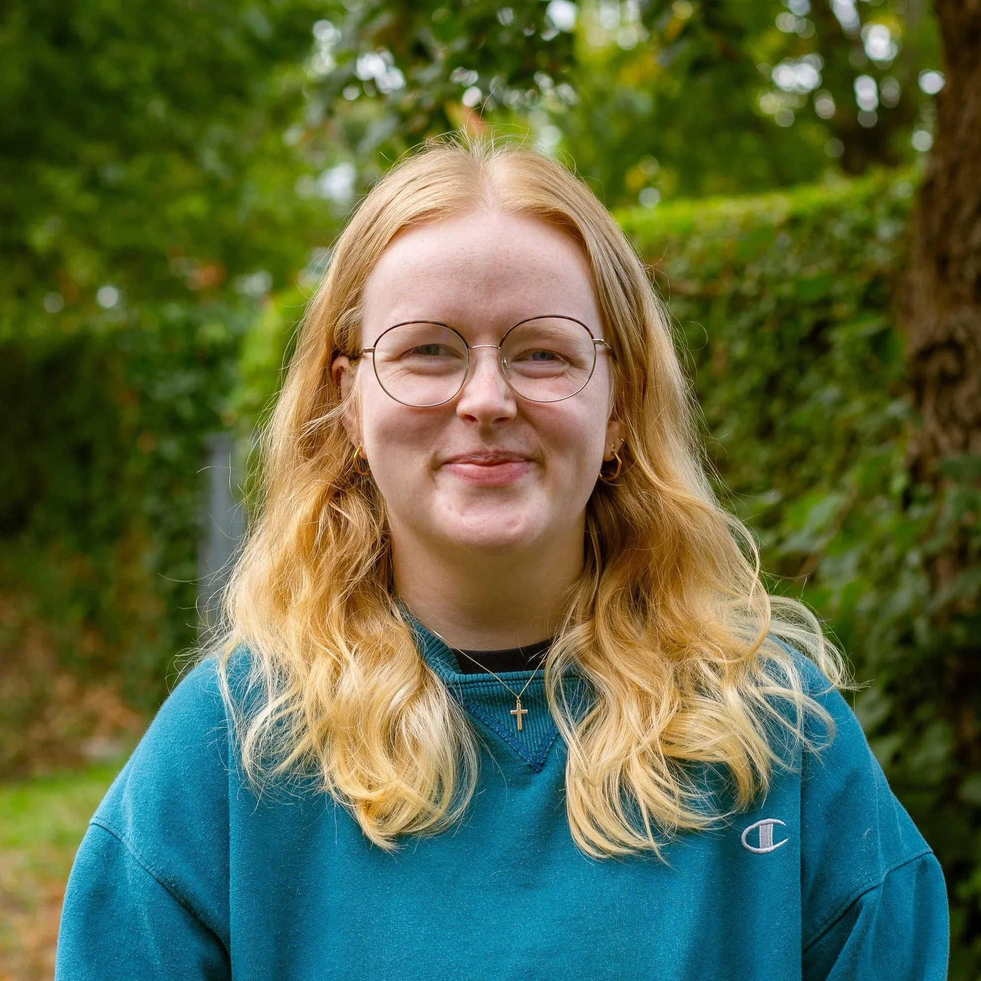 Young woman with red hair, glasses, and freckles smiling outdoors in front of green foliage and a brick wall.