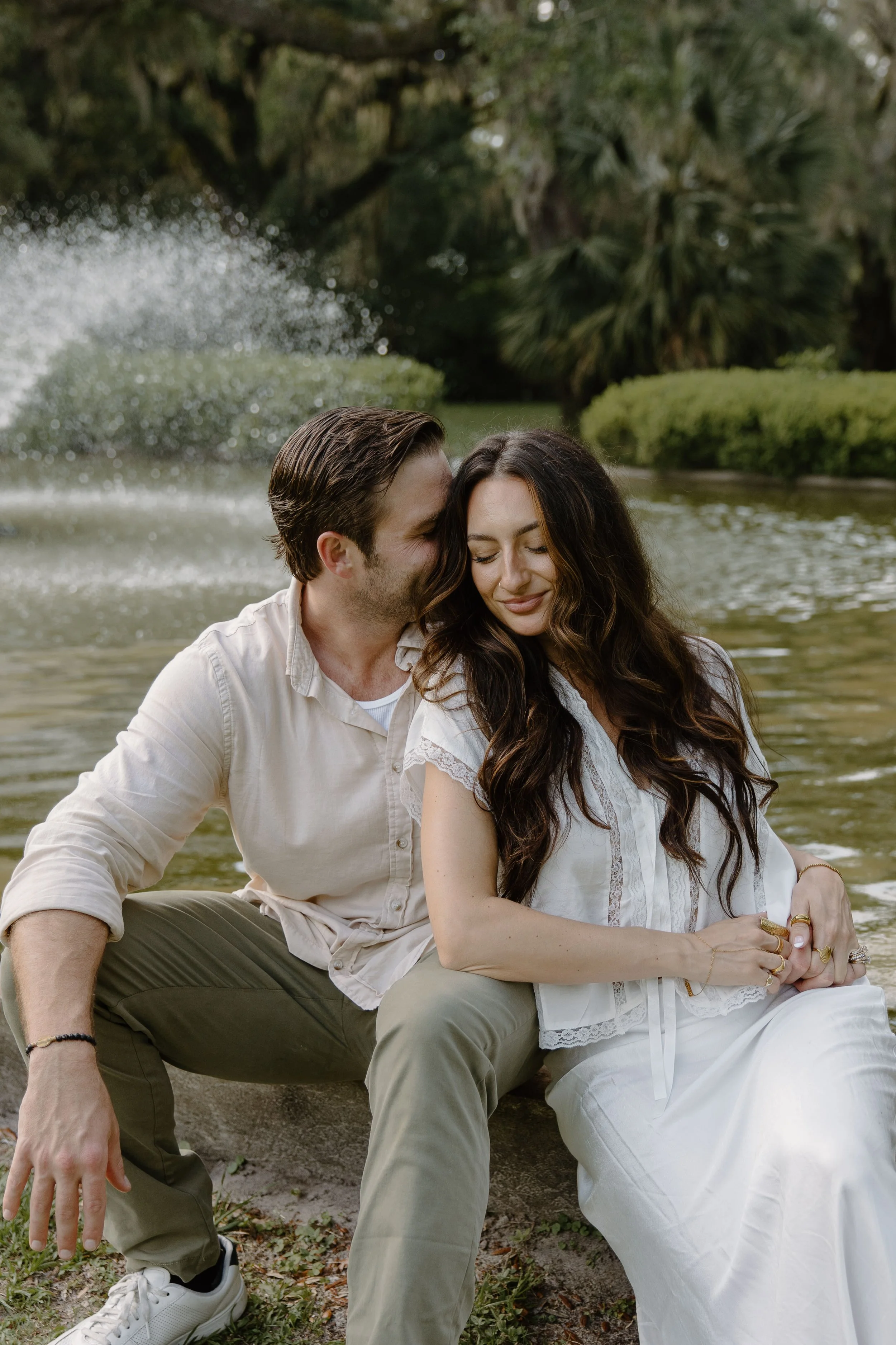 Meg Nailling sitting with her husband beside a pond and fountain surrounded by trees and greenery.