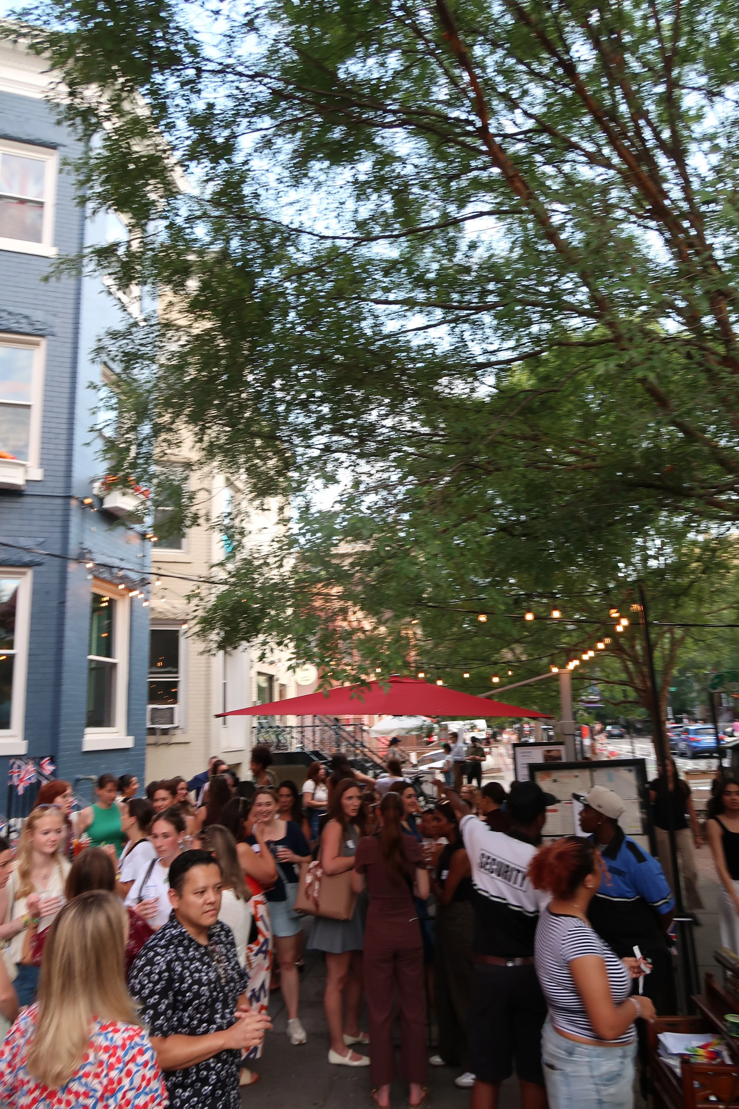 Crowd of women gathered outdoors at Meg Nailling’s DC Girls Meet Up at Duke’s Grocery in Dupont Circle, Washington, DC, beneath string lights and red patio umbrellas.