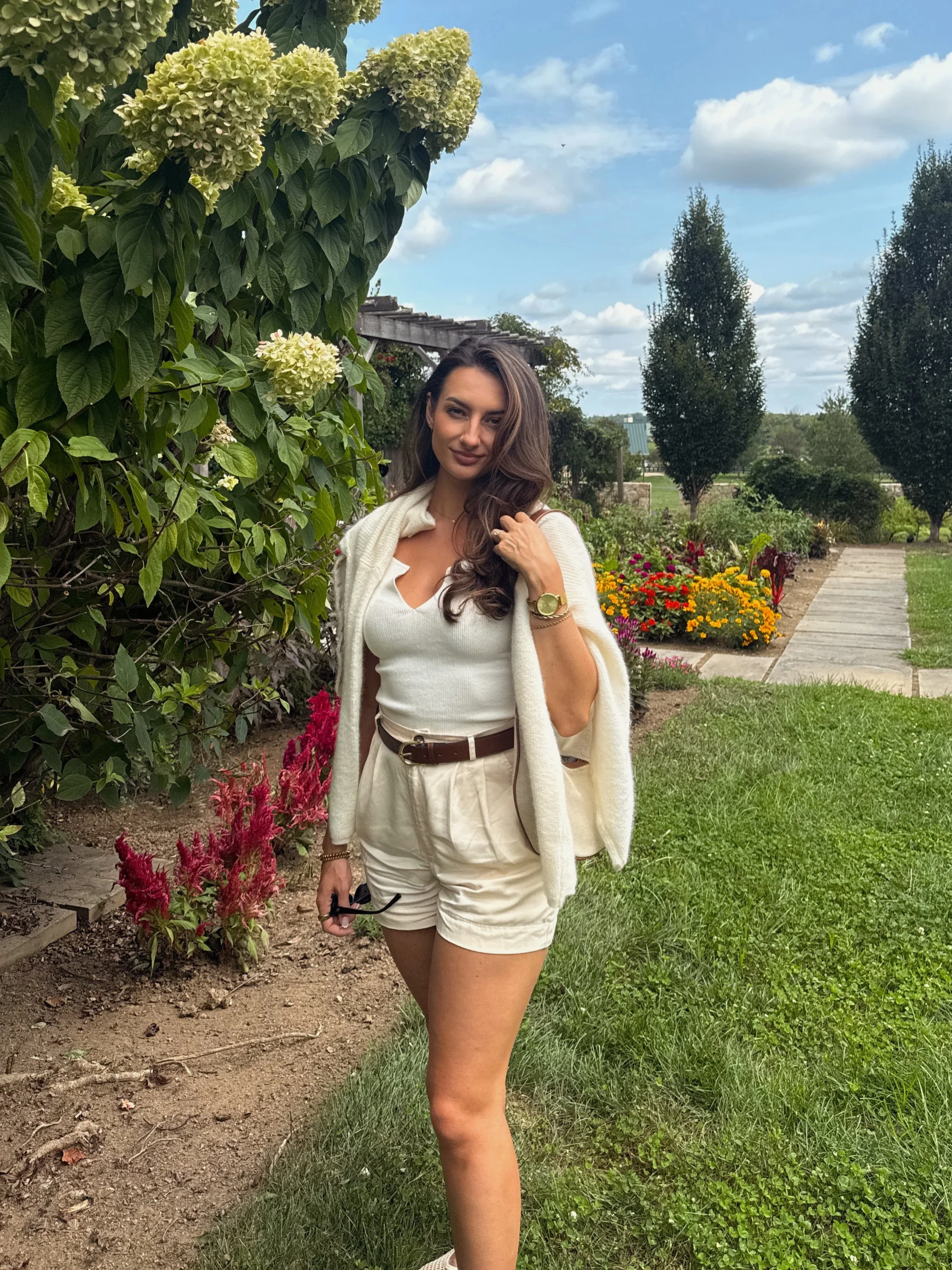 Meg Nailling wearing white shorts, a white top, and a beige cardigan while standing in a flower garden in Northern Virginia.
