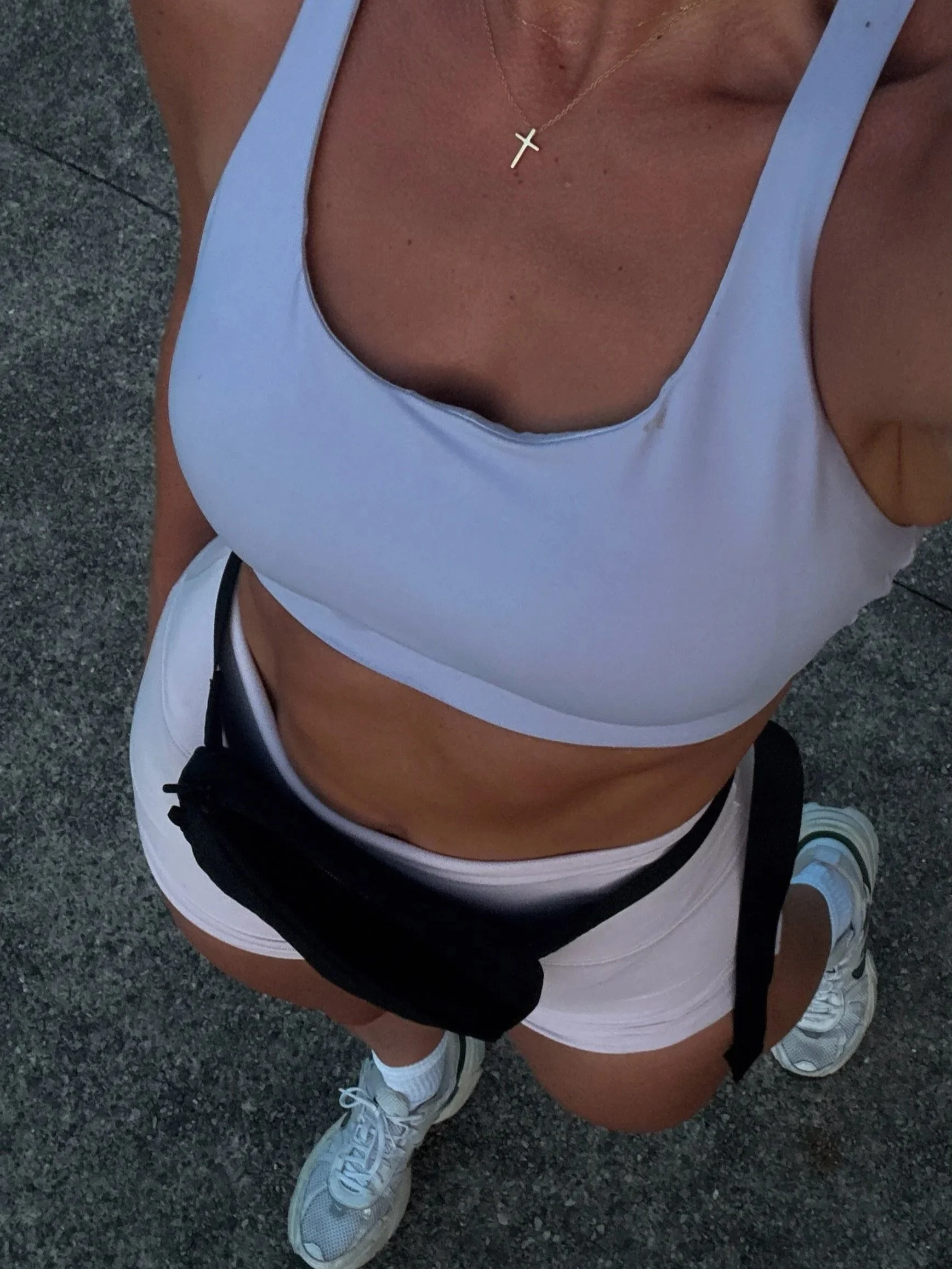 Meg Nailling in white athletic shorts, a white workout top, and sneakers, photographed from above while wearing a gold cross necklace.