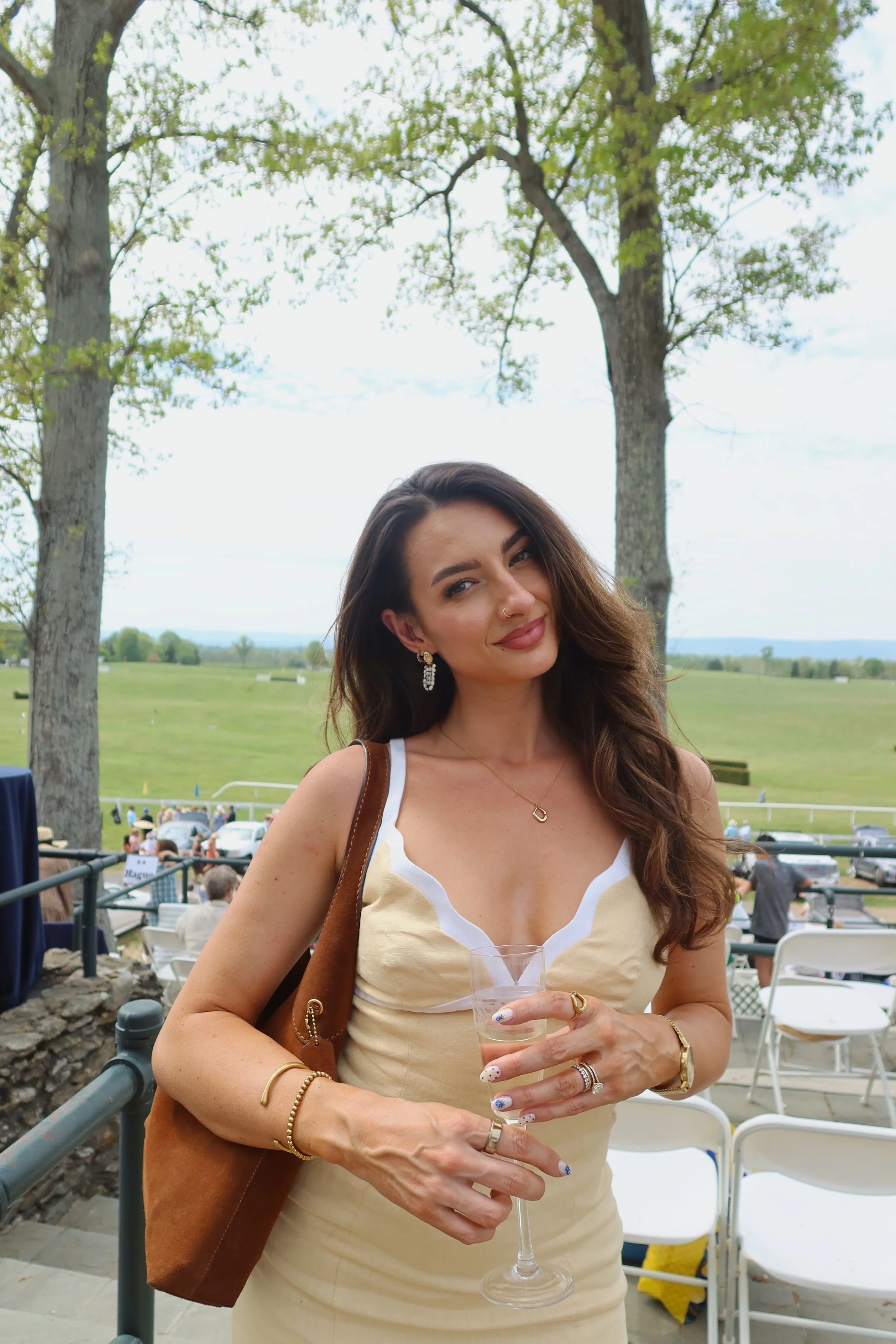 Meg Nailling wearing a cream-colored dress, gold jewelry, and a brown shoulder bag while holding a wine glass at an outdoor polo event in Northern Virginia.