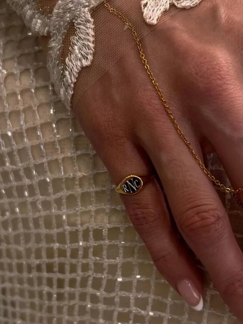 - Close-up of Meg Nailling’s hand wearing a gold signet ring and delicate gold bracelet against a textured cream fabric.