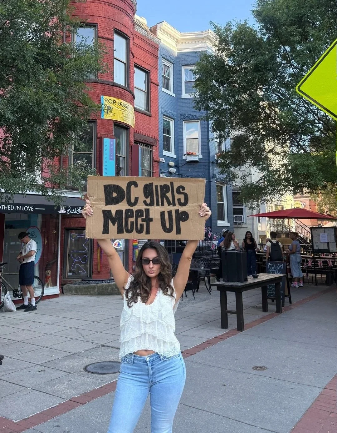 Meg Nailling holding a handmade “DC Girls Meet Up” sign on a sidewalk in Dupont Circle, Washington, DC, during a community event for women.