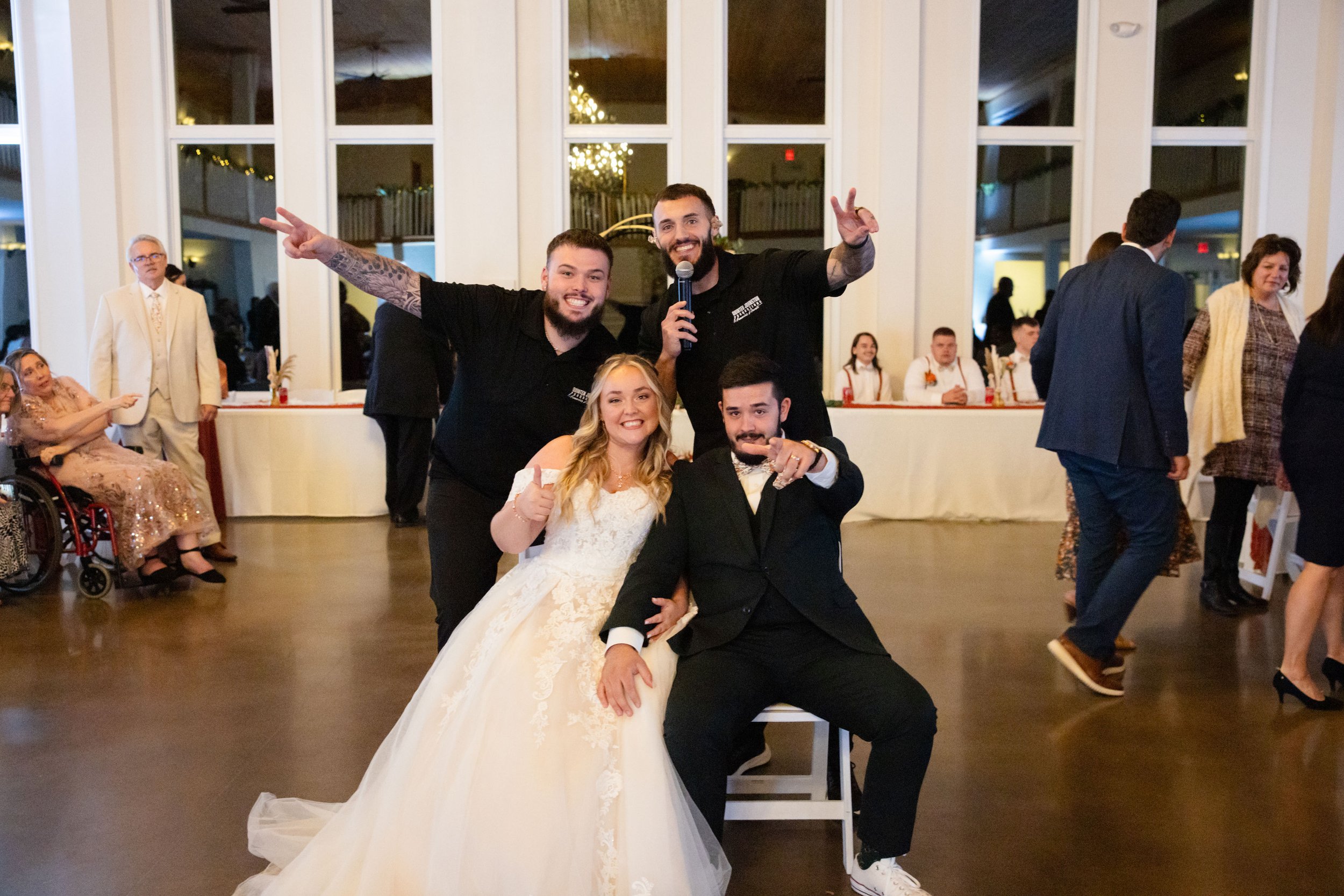 A wedding reception with a bride in a white wedding gown and a groom in a black tuxedo, sitting and posing with three men standing behind them, two of whom are making peace signs and one holding a microphone. Guests are visible in the background at t