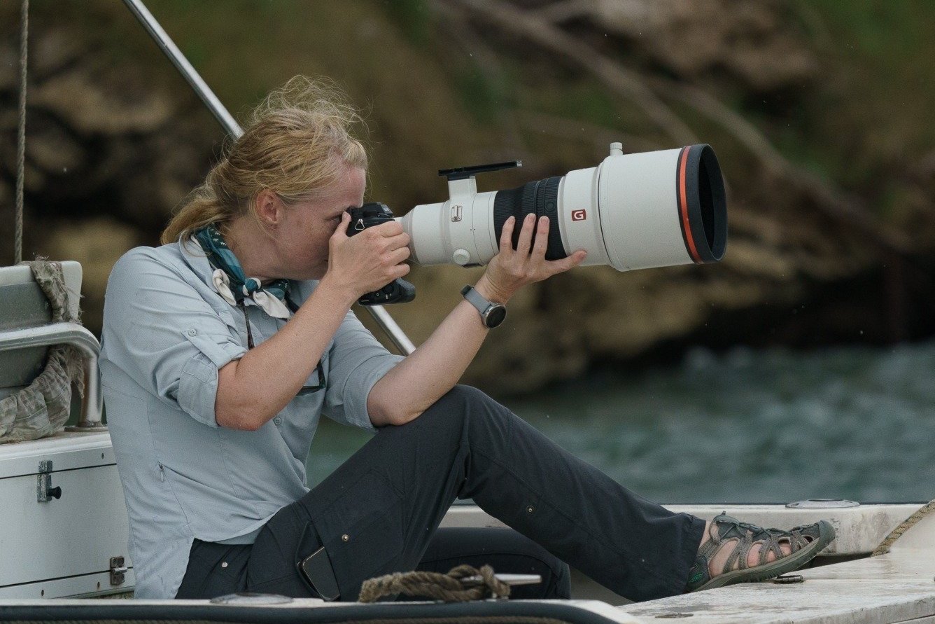 Annalise Kaylor sitting on a boat, looking through a large telephoto camera lens. She is outdoors near water with a rocky background.