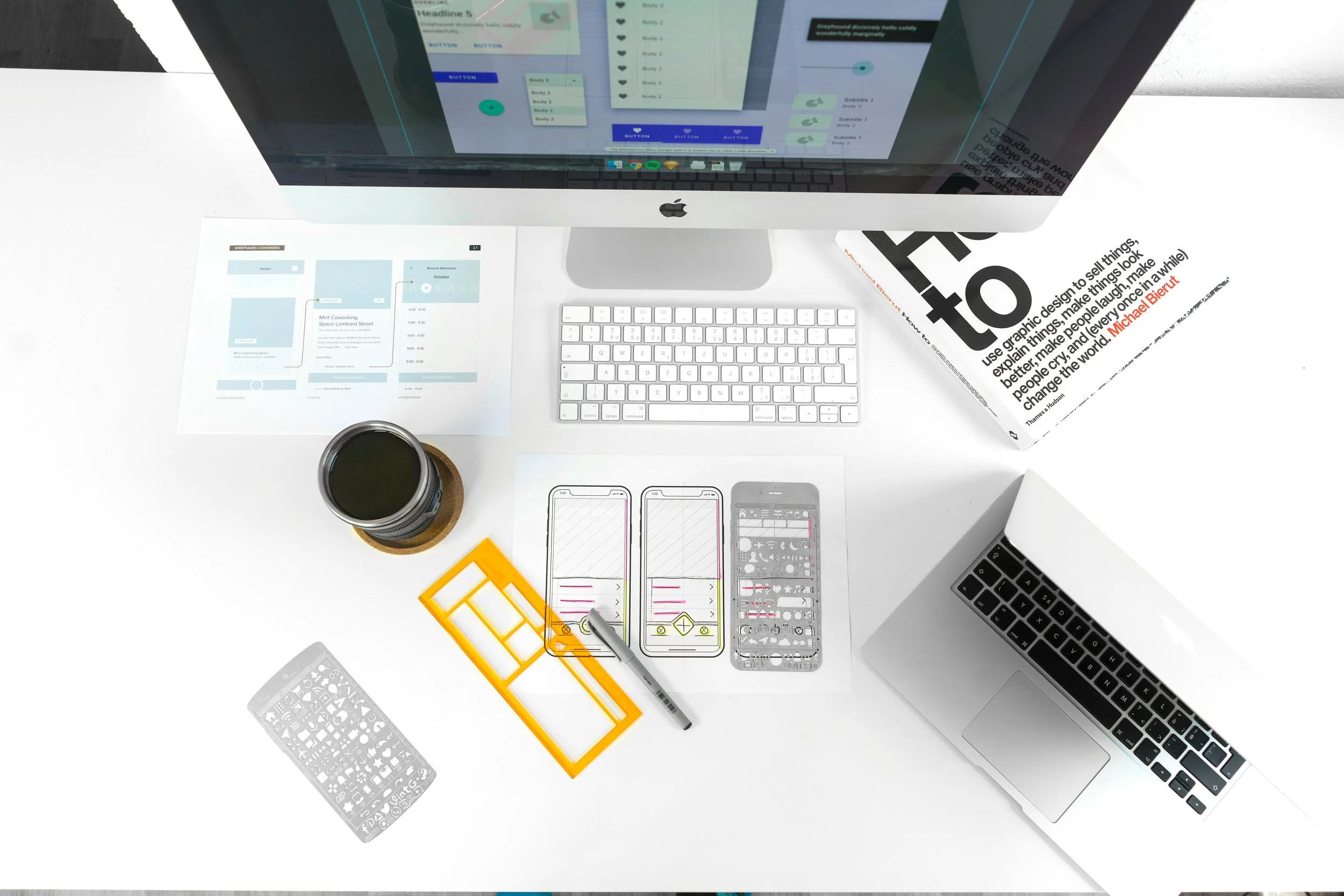 Overhead view of a work desk with a large iMac displaying design software, a white keyboard, a smartphone mockup, design templates, alphabets, a marker, a black coffee cup, and several sheets of paper, some with printed layouts and one with a quote about graphic design.