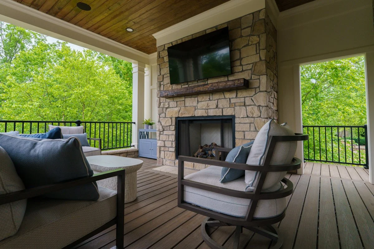 A cozy outdoor living space with a stone fireplace, a mounted flat-screen TV, and comfortable seating, overlooking green trees.