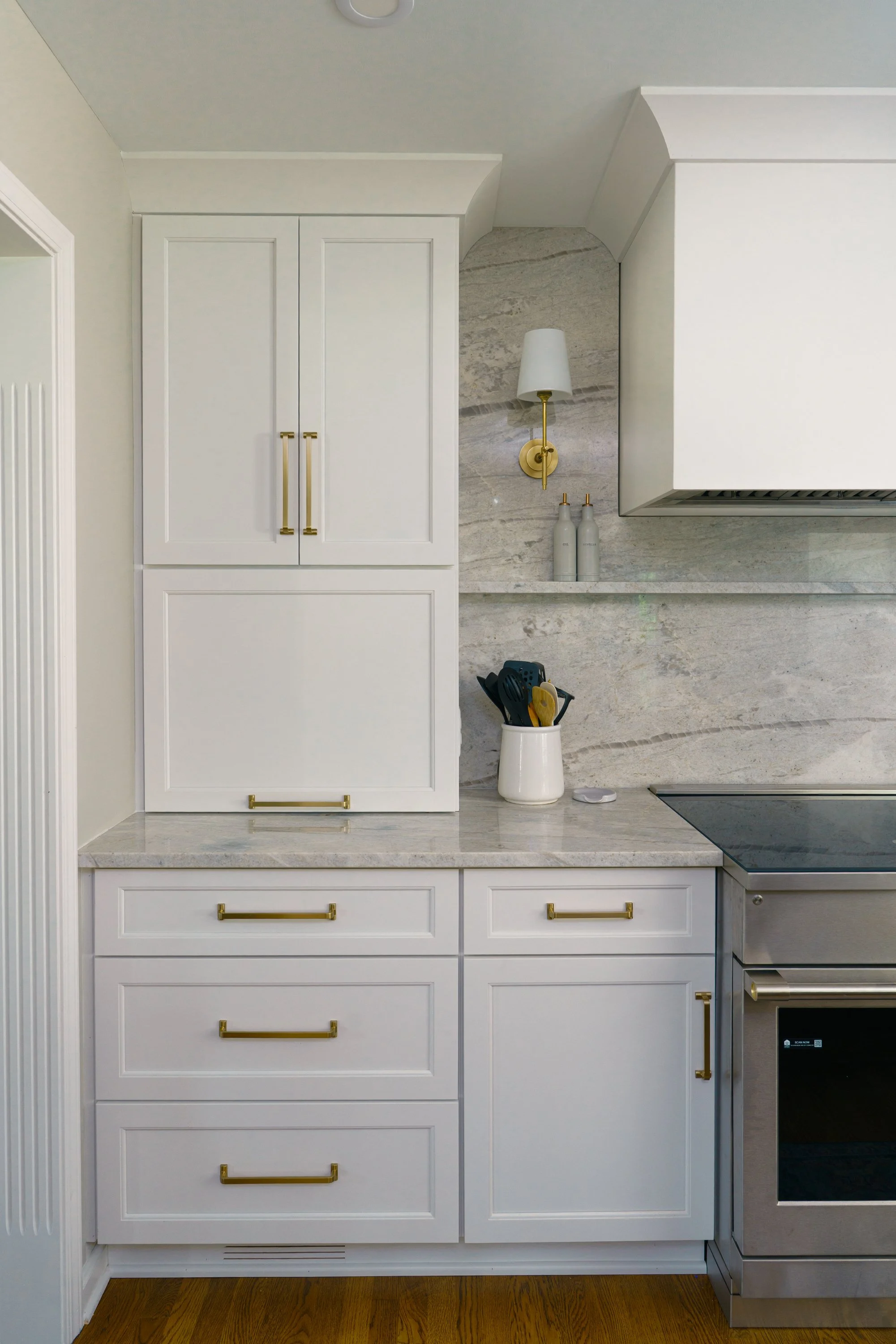 White kitchen cabinets with gold handles, a marble countertop, a wall-mounted lamp, and a countertop utensil holder with kitchen tools.