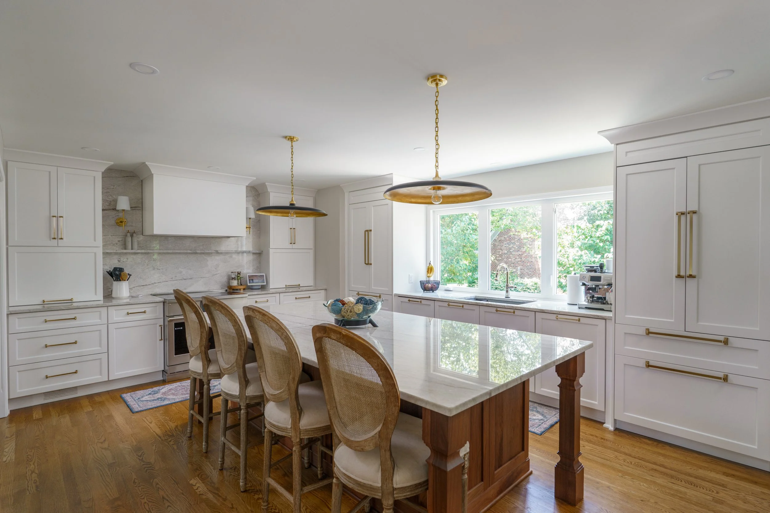 Bright kitchen with white cabinets, marble countertops, and a large island with four beige upholstered chairs, hardwood flooring, and two black and gold pendant lights hanging from the ceiling.