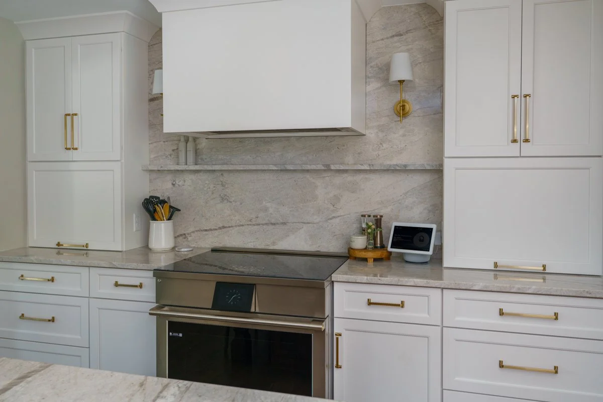Modern kitchen with white cabinets, gold handles, marble countertops, stainless steel oven, wall sconce, and a smart speaker on the counter.