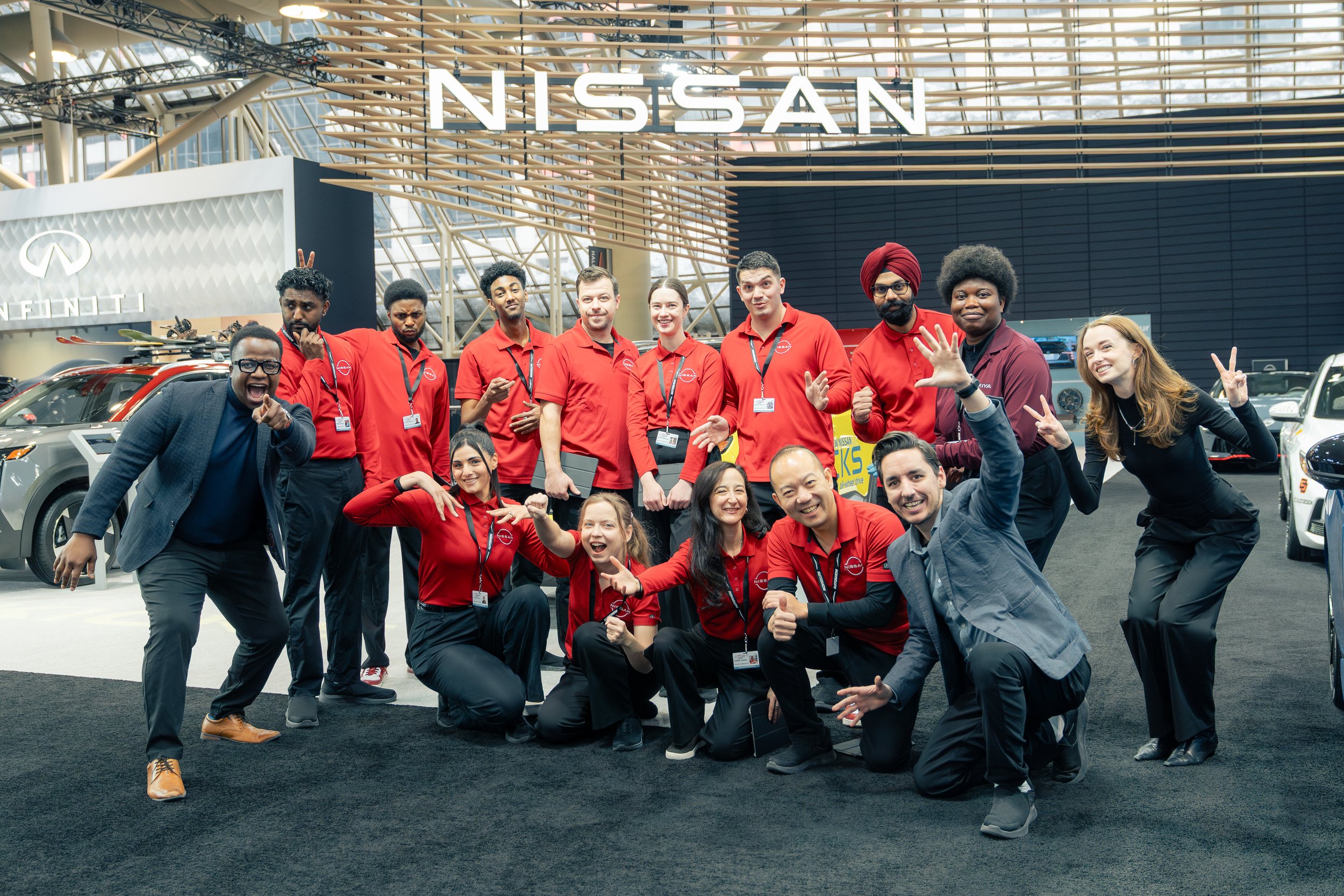 Group of Nissan employees posing at an auto show, all wearing red shirts with the Nissan logo and name tags, standing in front of a large Nissan display with a red background and logo.