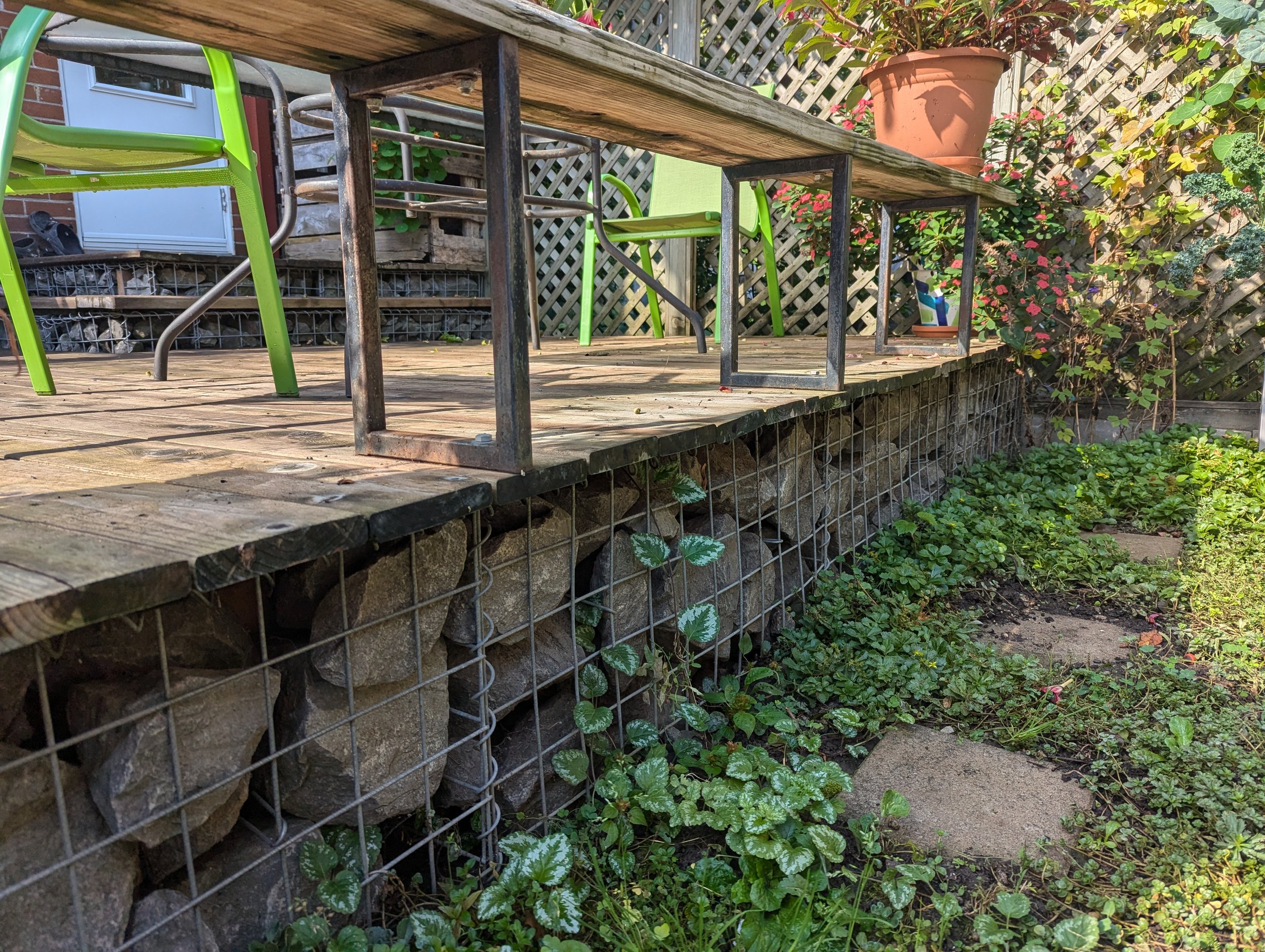 Terrasse en bois avec des chaises en plastique vert et un pot de fleurs, entourée de végétation et d'une clôture en métal.