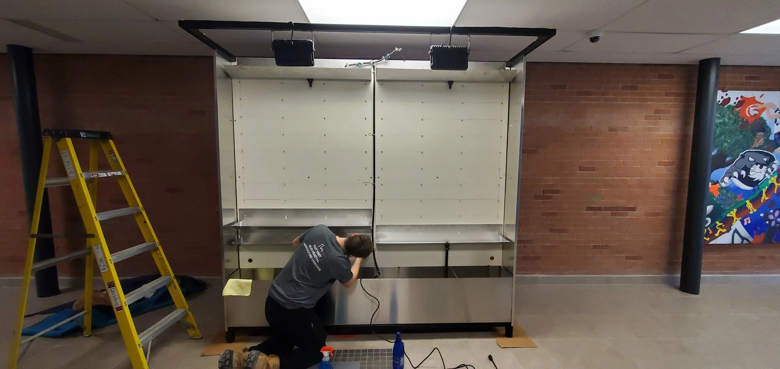Une personne en train d'installer ou de réparer un comptoir en stainless steel dans un espace intérieur avec un mur en briques rouges et une fresque murale colorée. Il y a une échelle jaune à gauche.