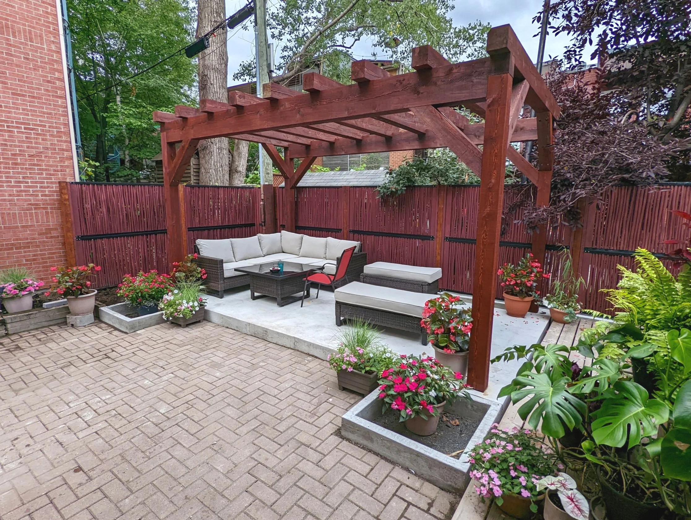 Un espace extérieur avec un coin salon comprenant un canapé d'angle, une chaise et un banc, sous une structure en bois, entouré de plantes en pot et d'une clôture en bois.