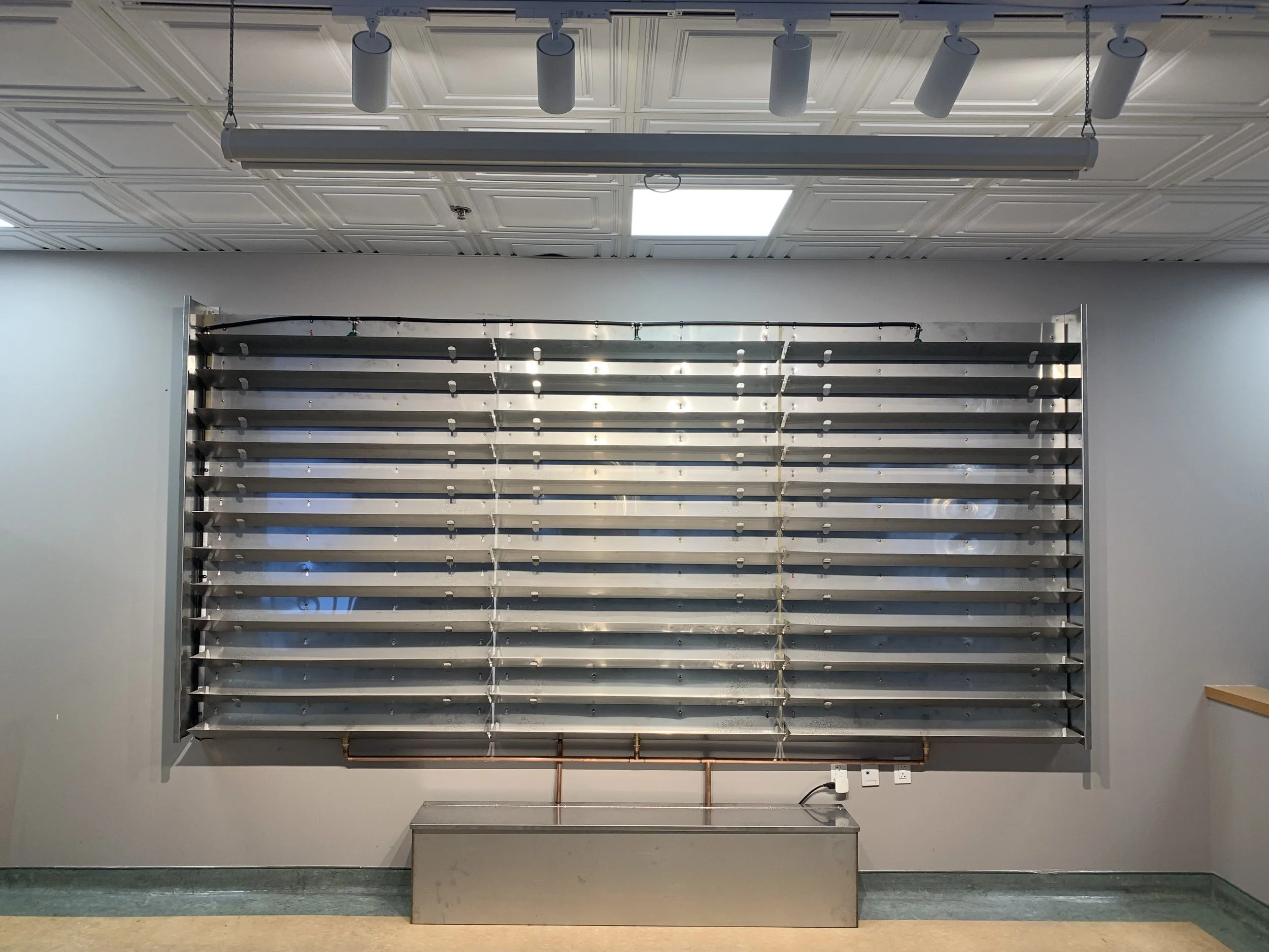 Une grande structure métallique avec plusieurs diagonalements, probablement un radiateur ou un dispositif de ventilation, fixé sur un mur dans une pièce lumineuse avec plafond en dalles.