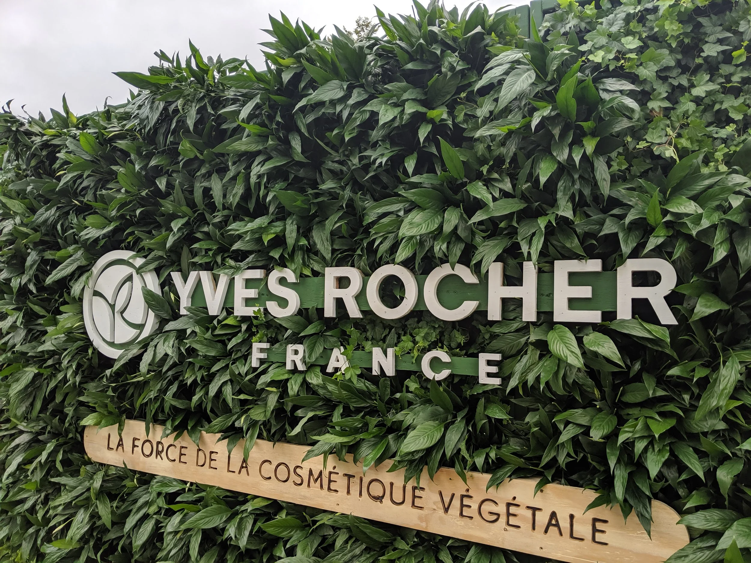Panneau indiquant 'Yves Rocher France' avec un logo stylisé de la marque, sur un mur de verdure dense, sous-titré 'La force de la cosmétique végétale' sur une planche en bois.