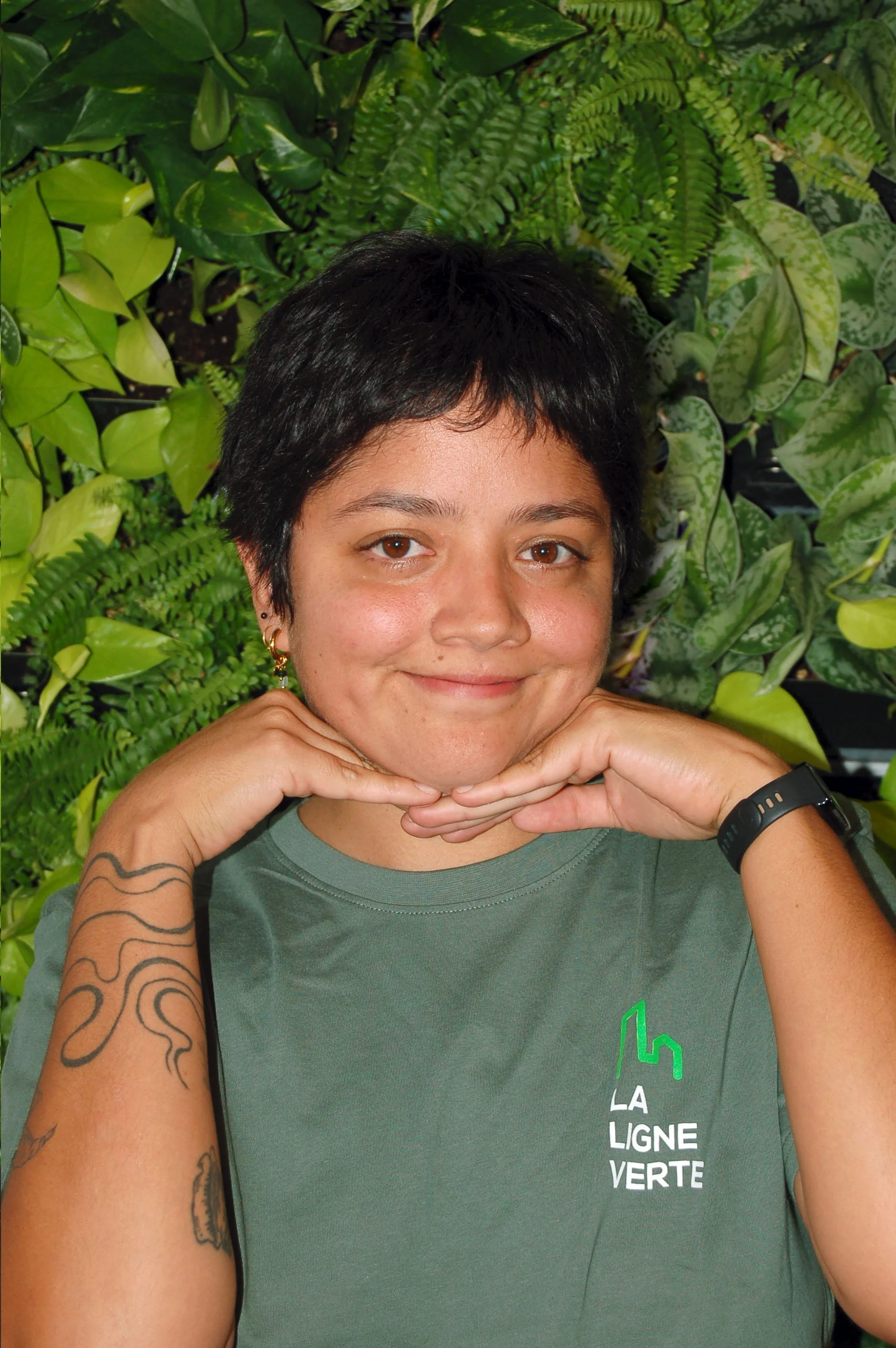 Portrait d'une personne souriante, portant un t-shirt vert avec le logo 'LA LIGNE VERTE', debout devant un mur recouvert de diverses plantes vertes.