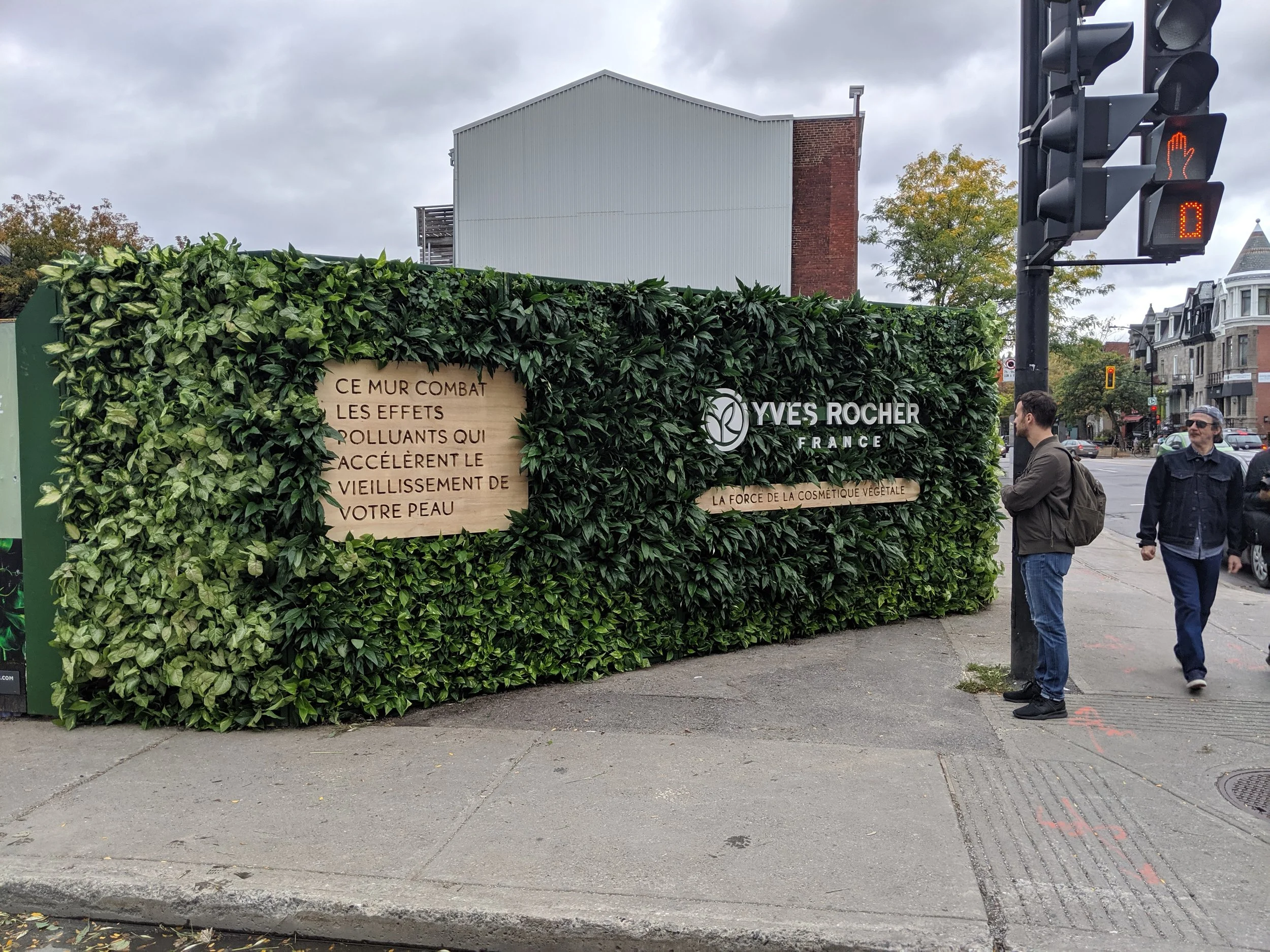 Panneau en bois derrière une clôture végétale avec le logo Yves Rocher et une inscription en français sur le thème cosmétique végétale, situé à un coin de rue. Des personnes attendent au passage piéton sous un ciel nuageux.