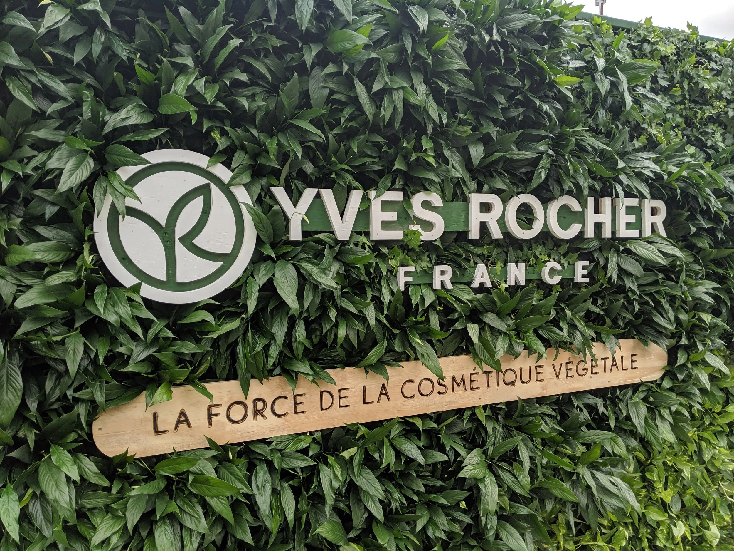 Plaque verte avec écritures blanches : 'Yves Rocher France' et un logo de feuille stylisée, entourée de feuillage. Bandeau en bois avec l'inscription : 'La force de la cosmétique végétale'.