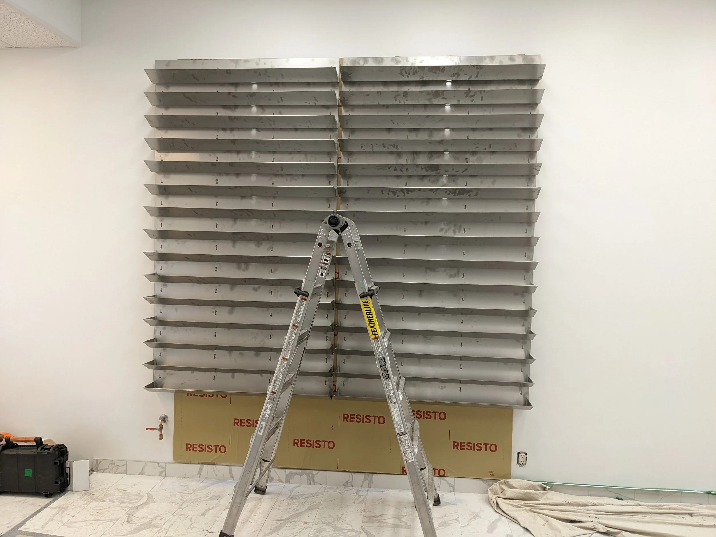 Mur en cours de rénovation avec un panneau de résistant en dessous, un escabeau en Aluminium en position centrale, un tuyau d'eau, un sac en toile et une boîte à outils sur le sol.