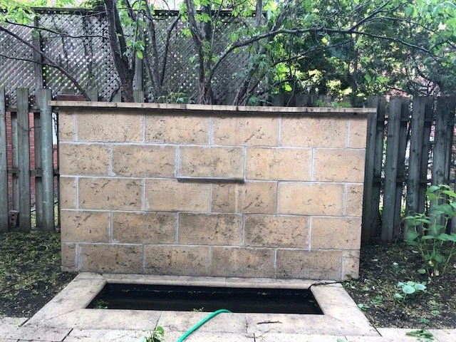 Un mur en blocs de pierre avec une cheminée ou un feu de camp en dessous, une corde verte accrochée et un terrain extérieur avec une clôture en bois et des arbres.