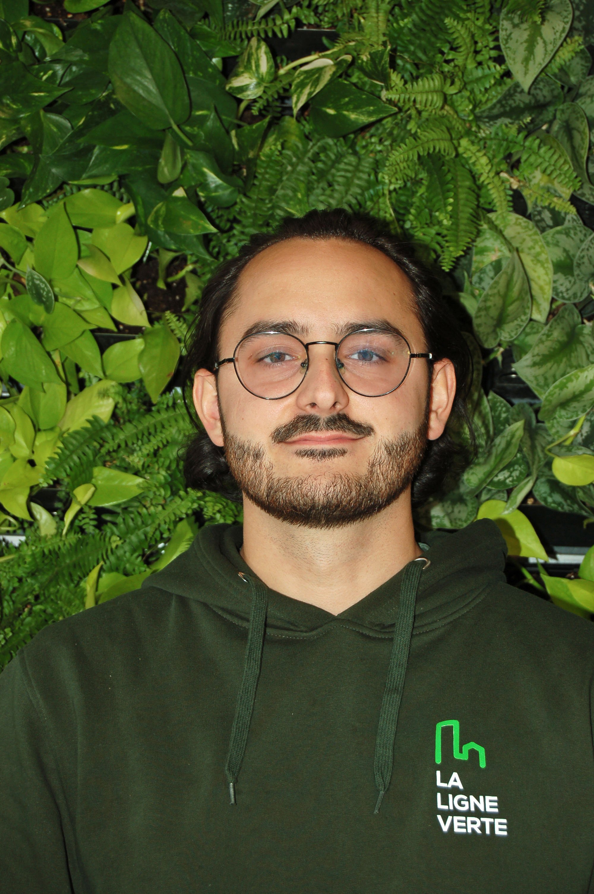 Un homme portant un sweat à capuche vert avec le logo de 'La Ligne Verte' devant un mur de plantes vertes.
