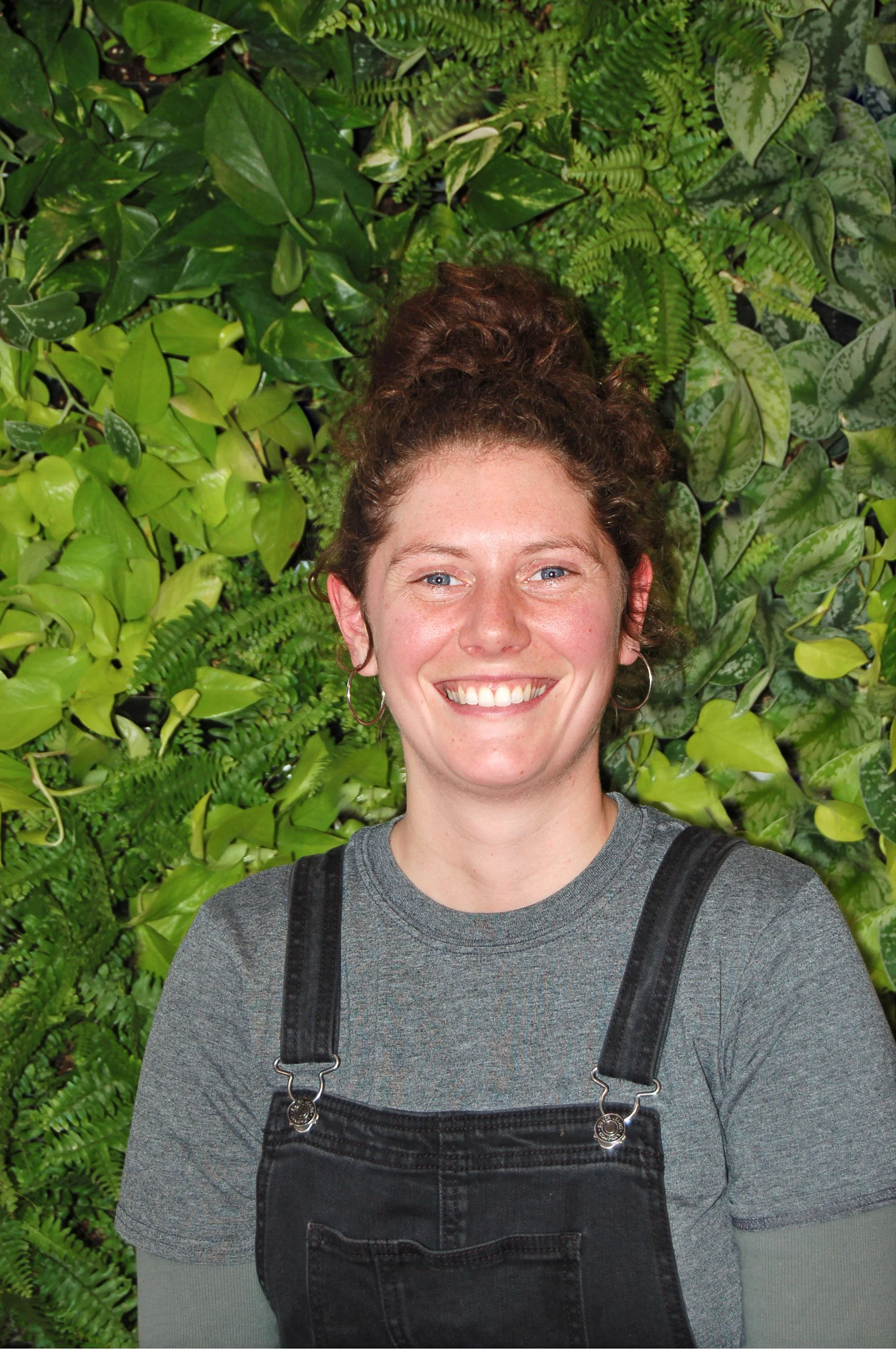 Jeune femme souriante avec des cheveux bouclés en chignon, portant un t-shirt gris et un salopette noire, devant un mur de plantes vertes.