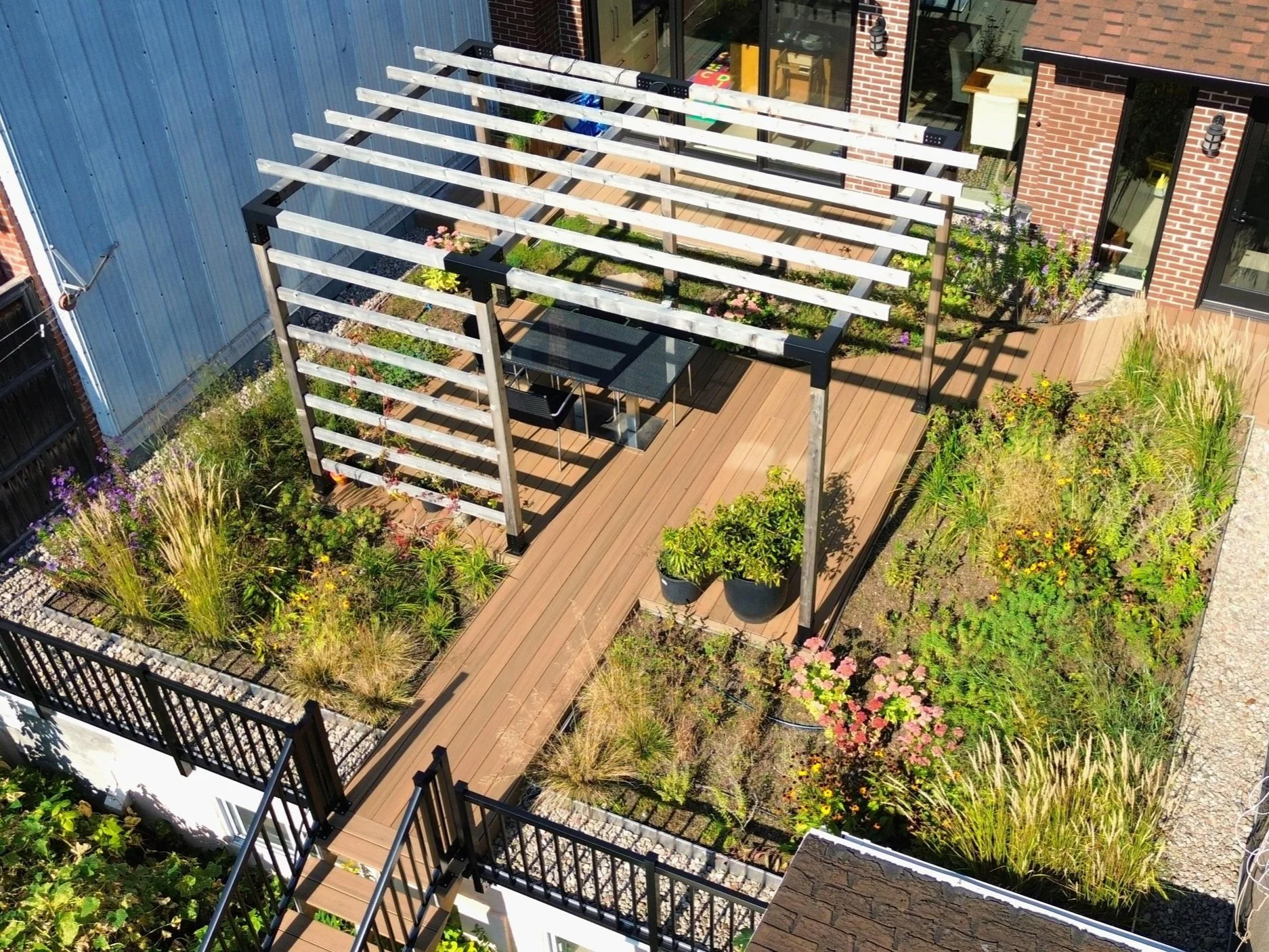 Jardin urbain avec pergola en bois, table et chaises, plantes et fleurs variées, balcon en bois, entouré de clôtures en métal.