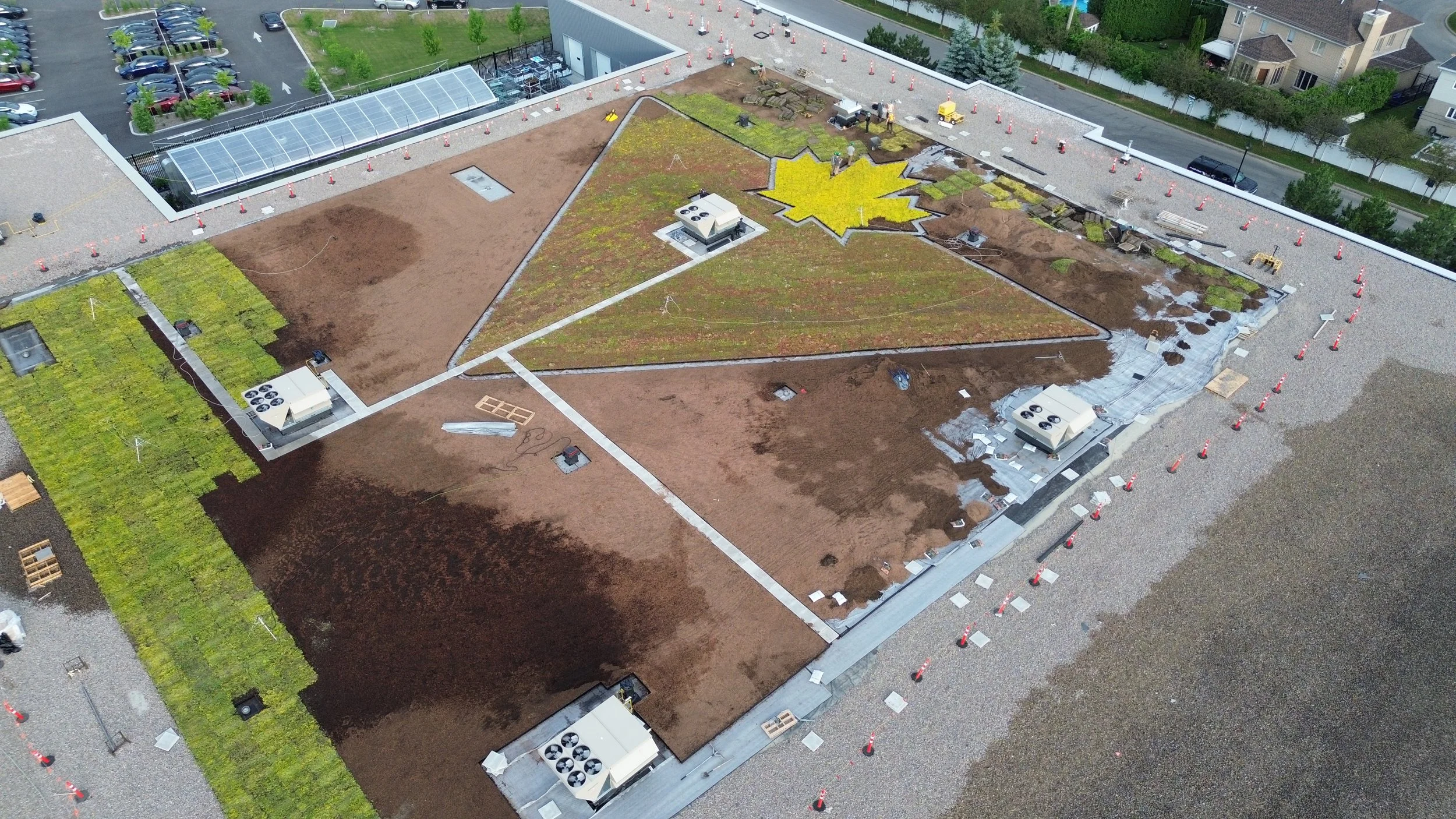 Travaux de toiture en cours sur un toit plat, avec différentes sections en cours d'aménagement, dont une avec un motif de feuille d'érable jaune. Des équipements techniques et des matériaux de construction sont visibles.