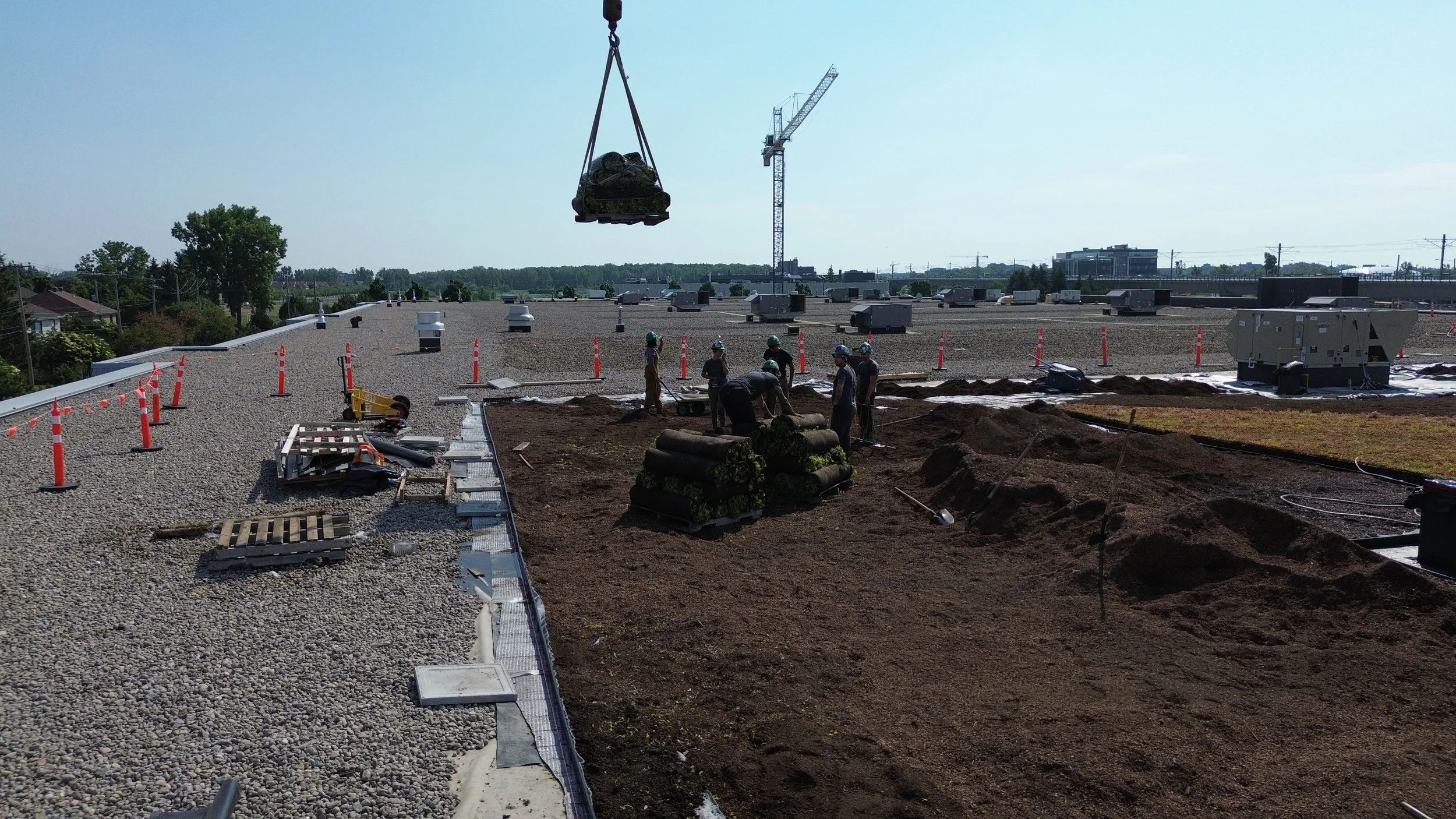 Travaux de construction sur un toit avec plusieurs ouvriers, matériaux, un grue soulevant des objets, et un ciel clair.